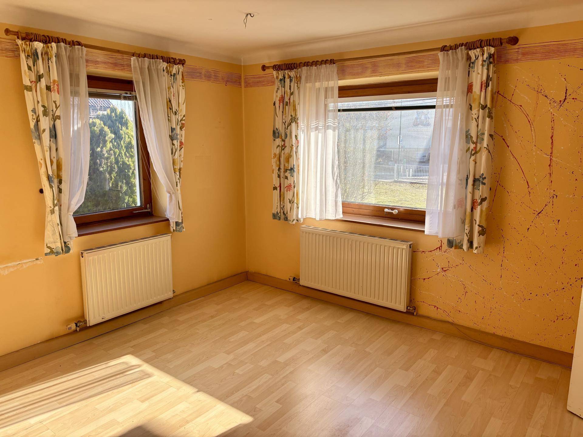 Helles Zimmer mit gelben Wänden, zwei Fenstern und Laminatboden, bereit zur Renovierung.