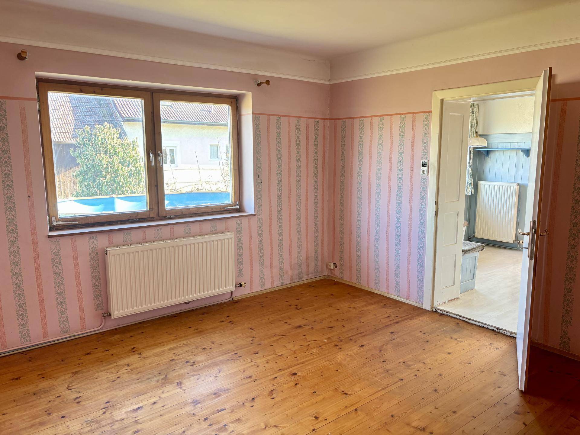 Geräumiges Zimmer mit rosa gestreifter Tapete, Holzdielenboden und Blick ins Grüne.