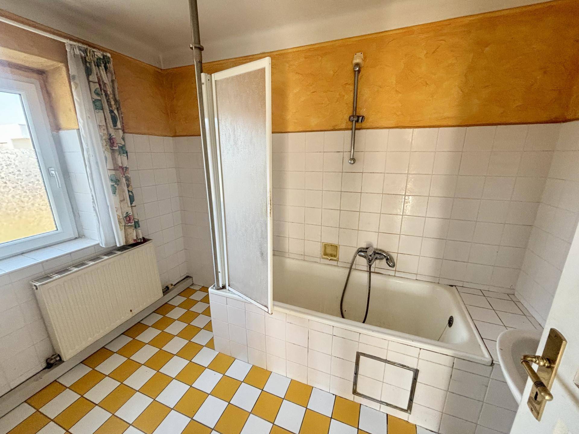 Badezimmer mit gelb-weißen Fliesen, Badewanne mit Duschfunktion und Fenster.
