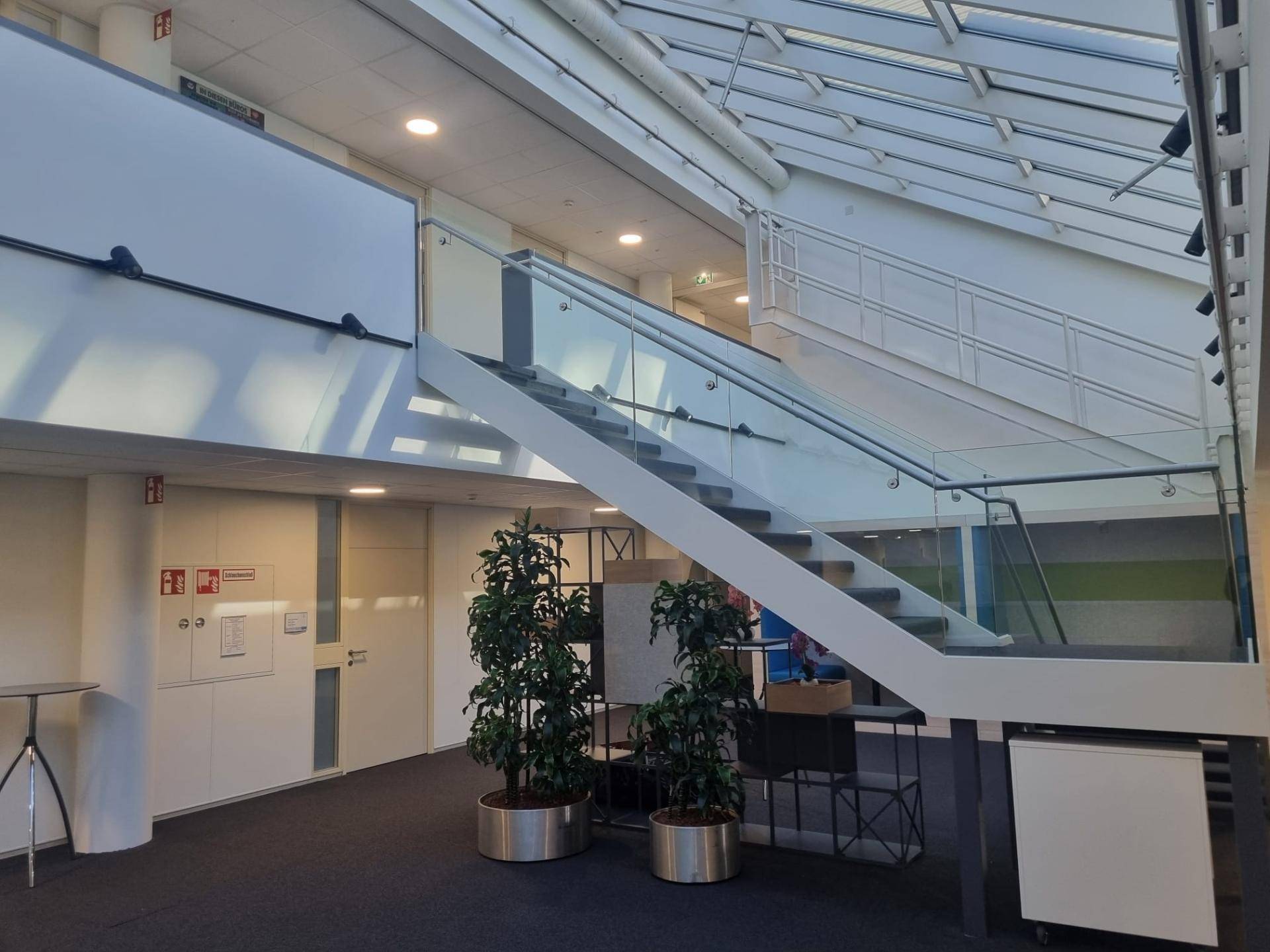 Helle Eingangshalle mit moderner Treppe, Glasgeländer und viel Tageslicht durch das beeindruckende Glasdach.
