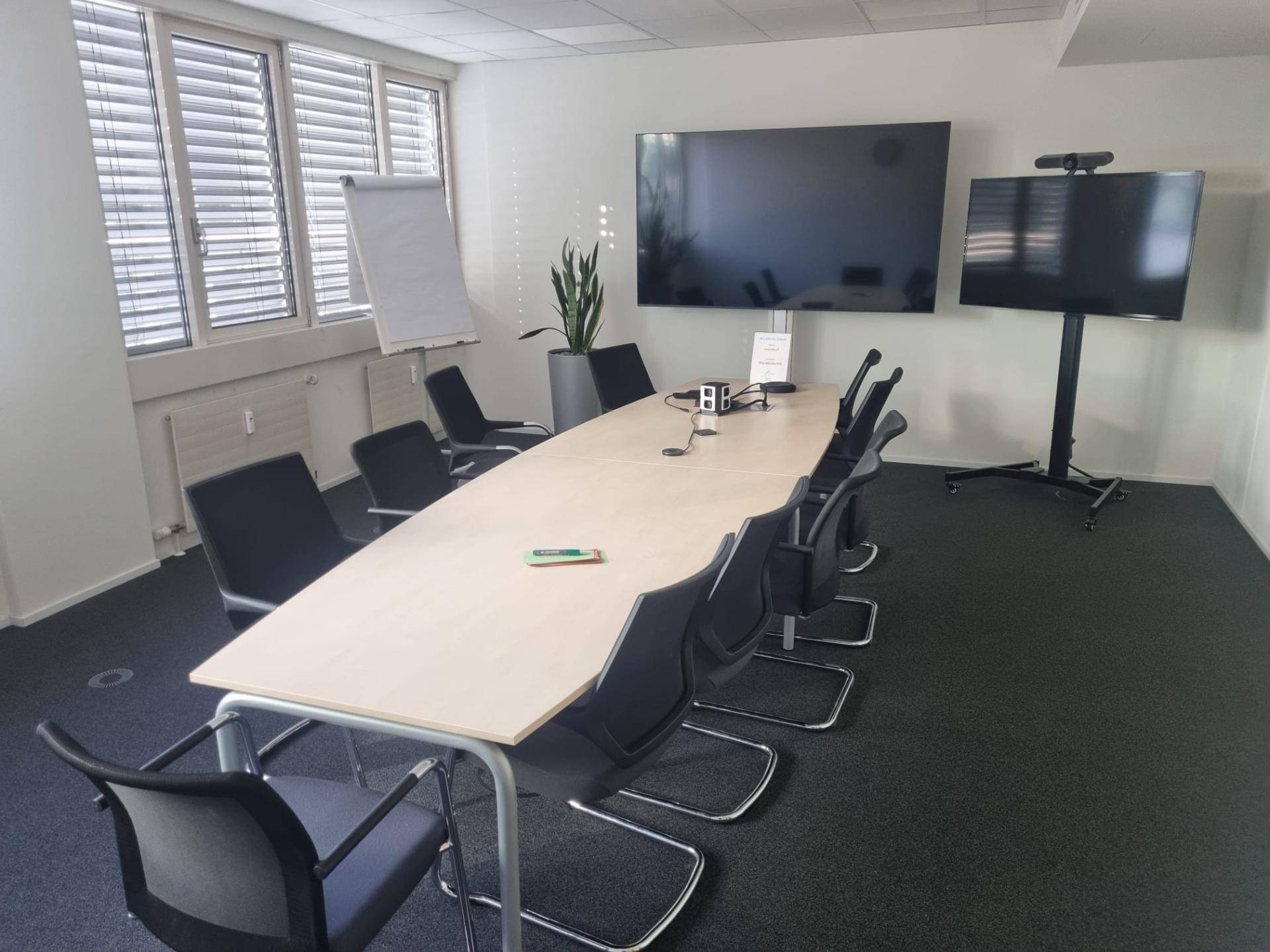 Moderner Konferenzraum mit großem Tisch, bequemen Stühlen und zwei Bildschirmen für Präsentationen, ideal für Besprechungen.