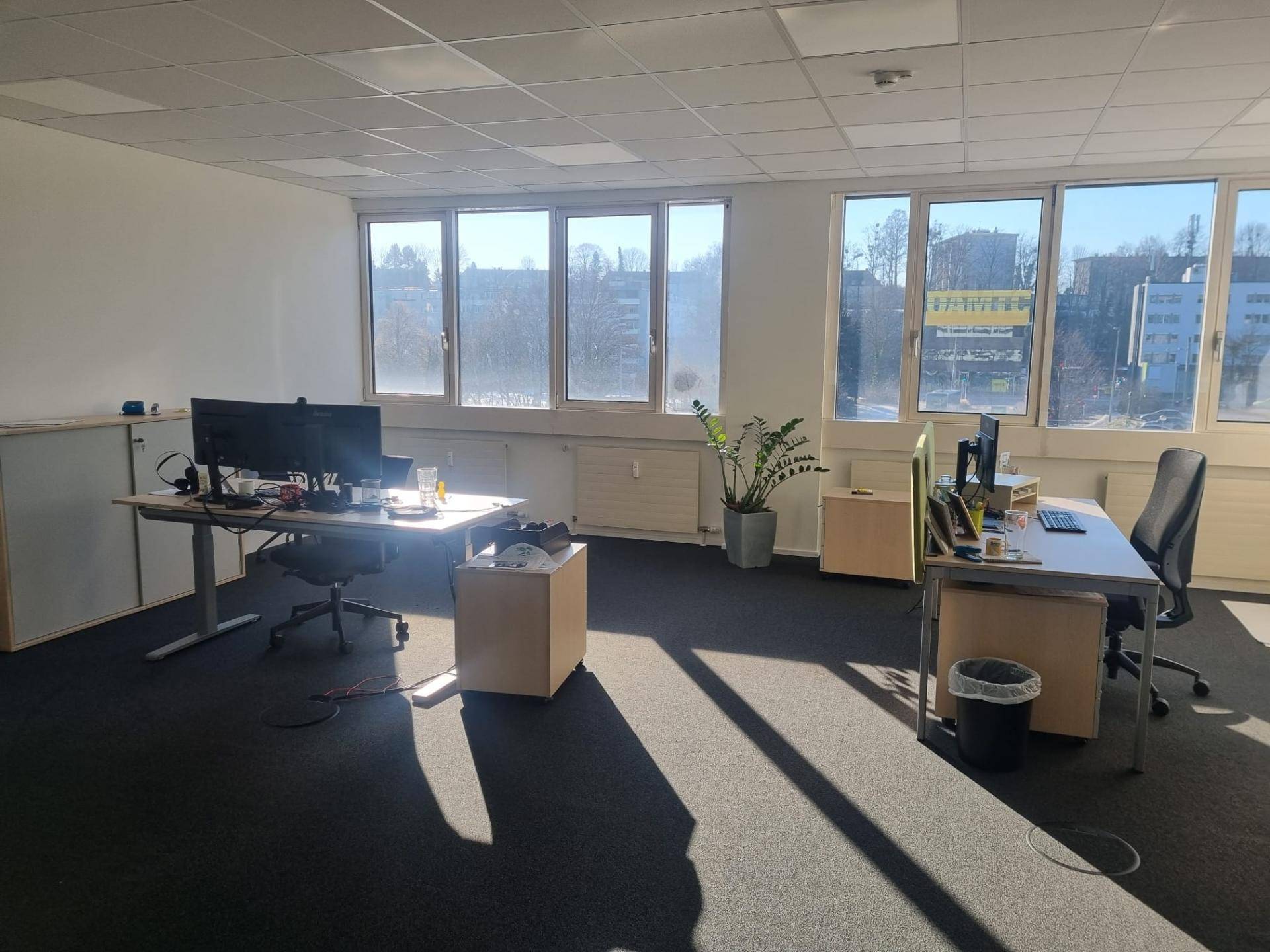 Helles Büro mit mehreren Arbeitsplätzen, großen Fenstern und Blick auf die grüne Umgebung, gut beleuchtet.