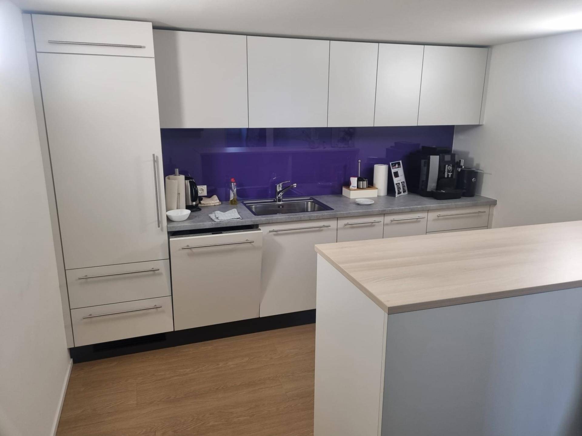 Moderne Teeküche mit weißen Schränken, lila Spritzschutz und Holzboden, gut ausgestattet für Mitarbeiter.