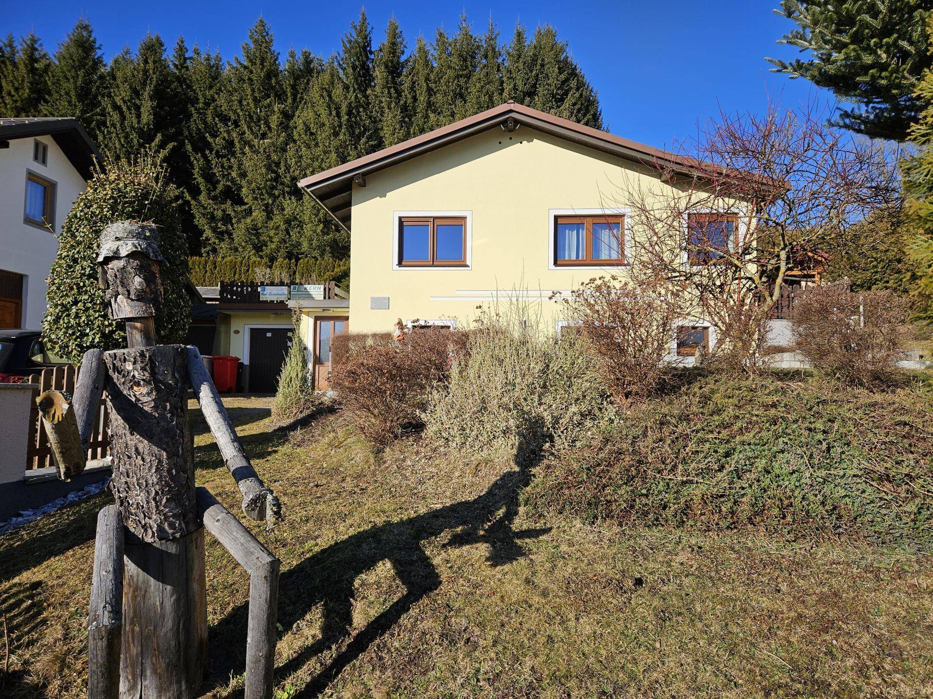 Gelbes Einfamilienhaus mit Garten und Baumbestand, gesehen von der Vorderseite mit Holzfigur.