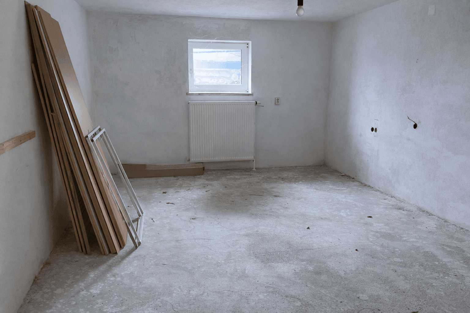 Unfertiger Raum mit Betonboden, Fenster und Baumaterialien an der Wand.
