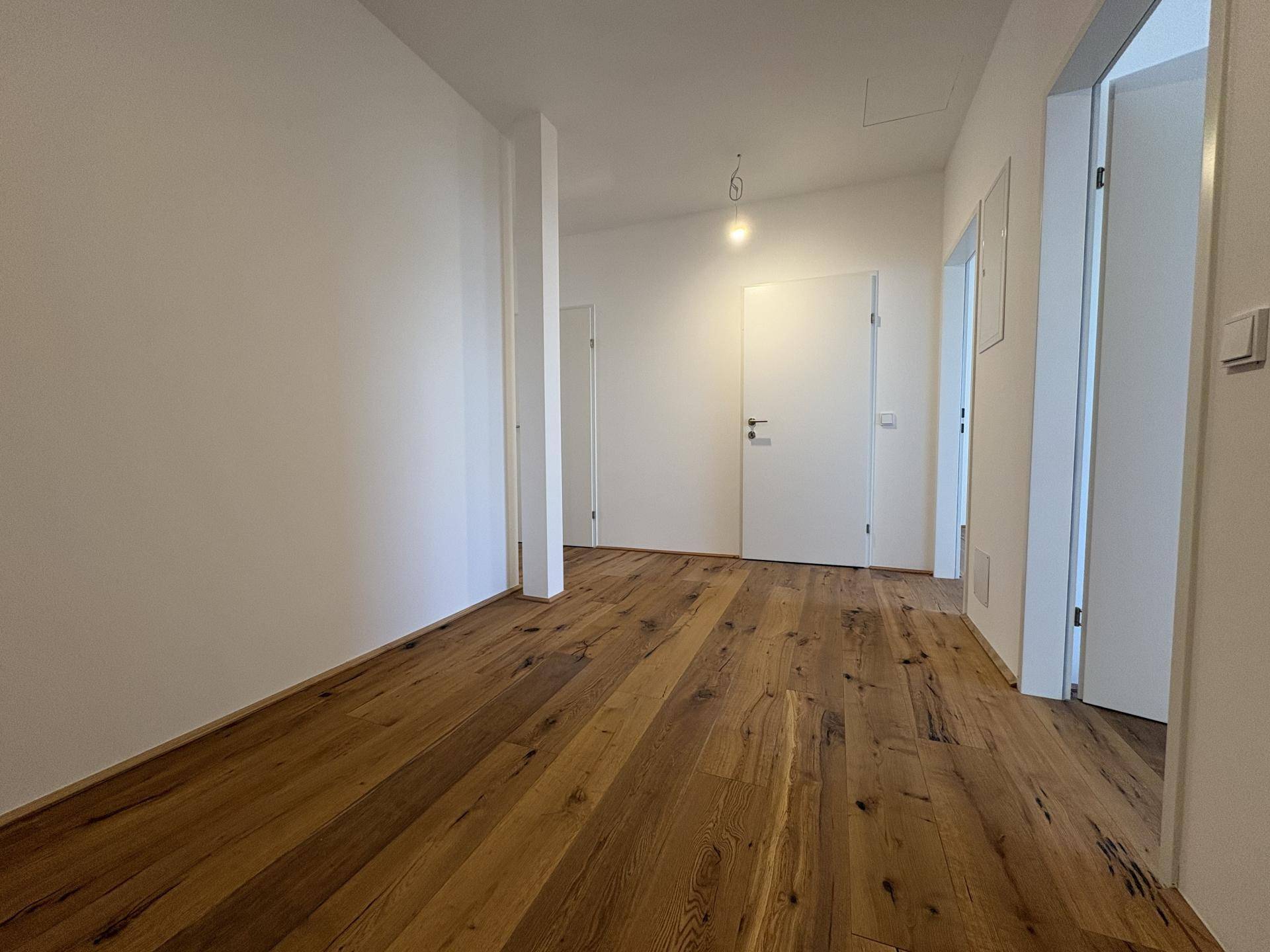 Langer Flur mit warmem Holzfußboden, weißen Wänden und mehreren Zimmertüren in einem Neubau.