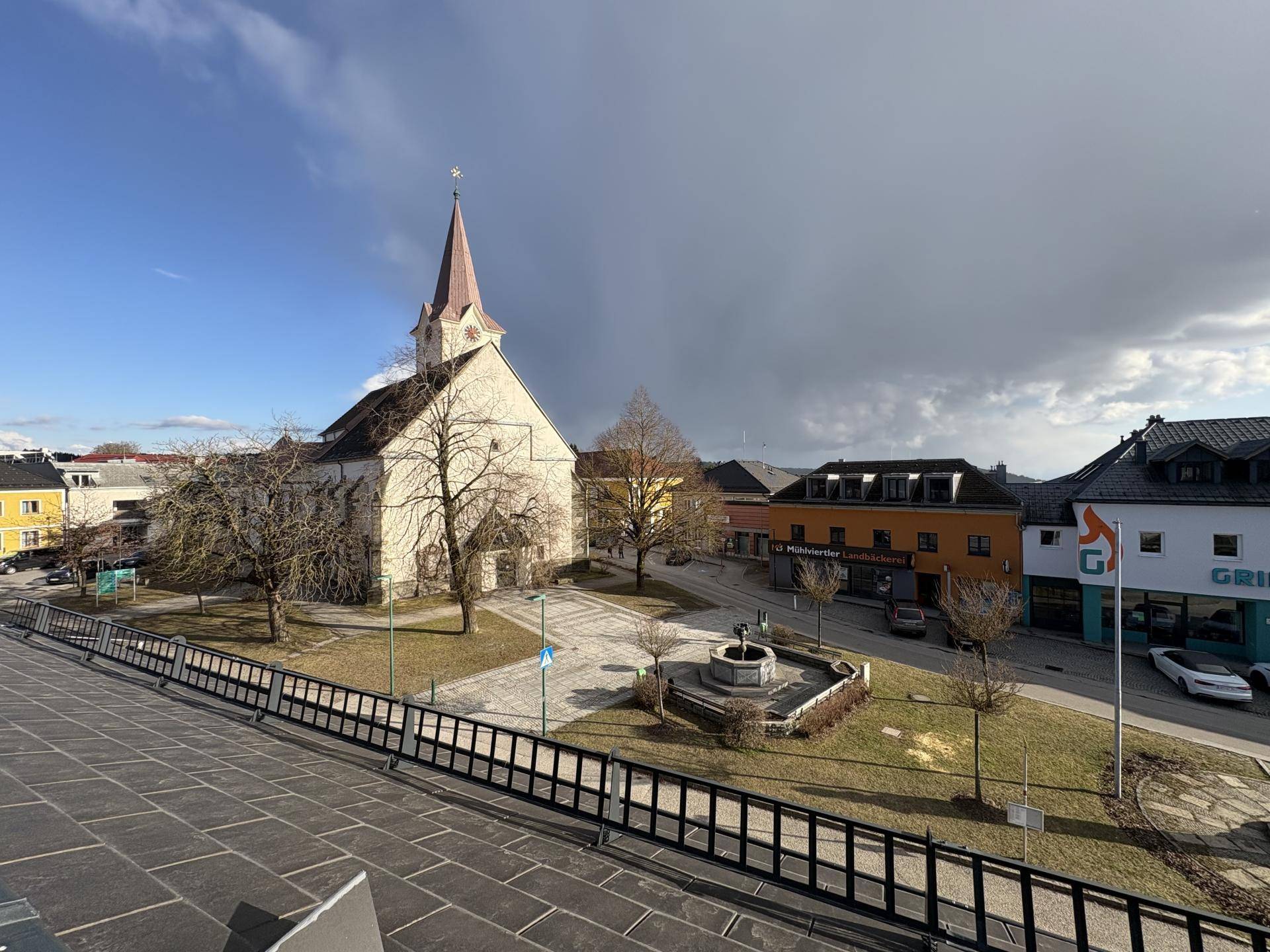 Weitläufiger Blick von der Terrasse auf den Stadtplatz mit Kirche, Bäumen und umliegenden Gebäuden.