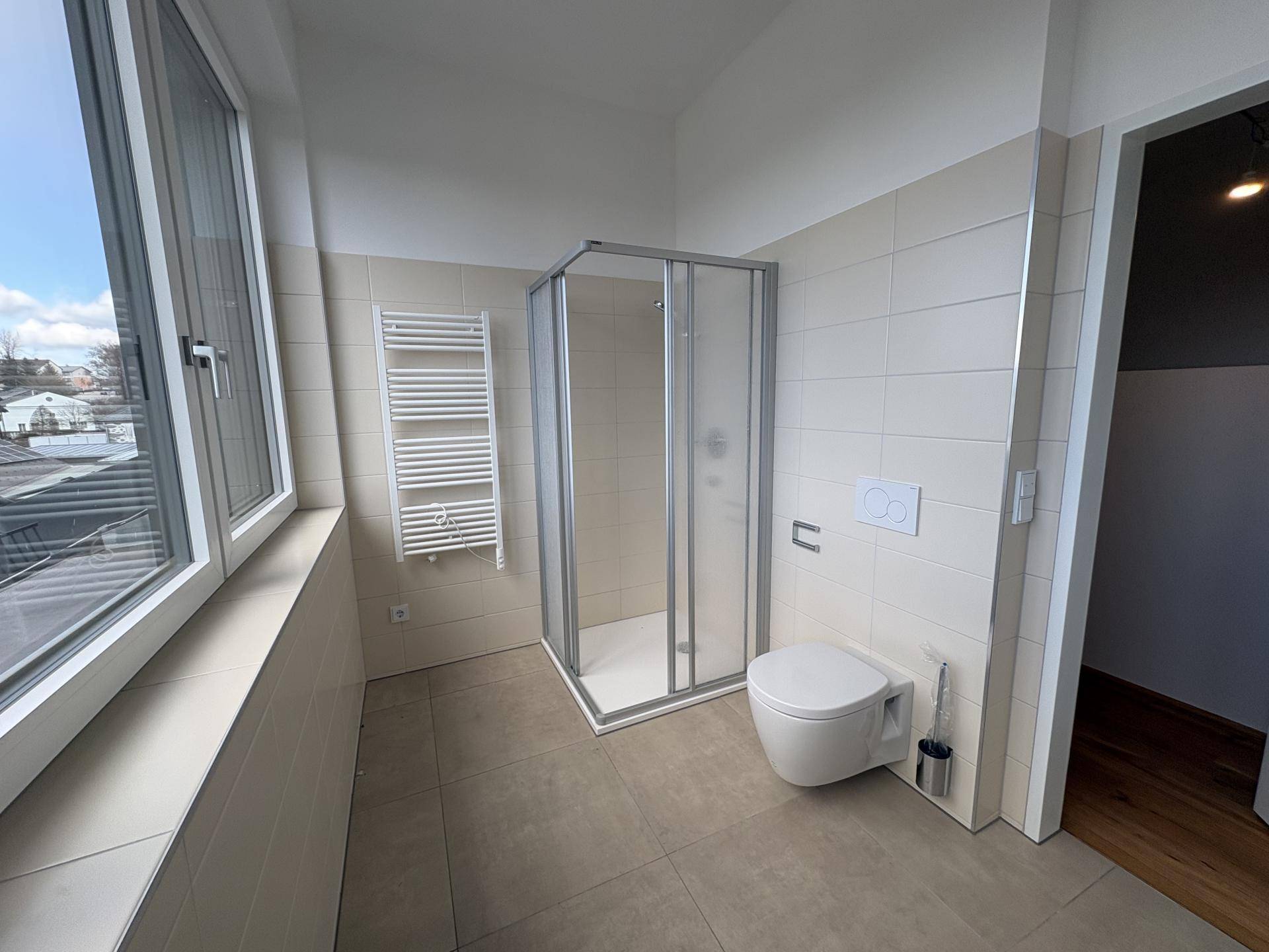 Helles Badezimmer mit Duschkabine, Wand-WC und großem Fenster, das viel Tageslicht hereinlässt.