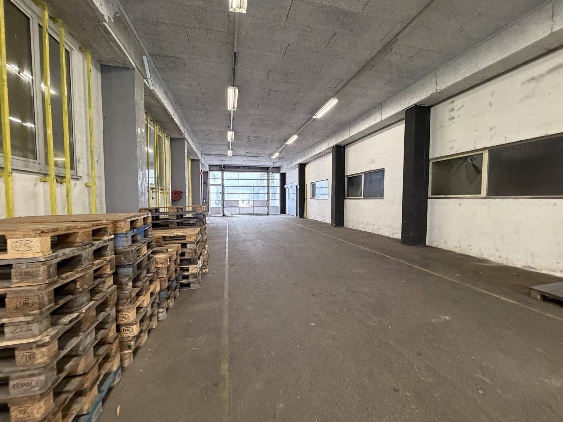 Langer Lagerraum mit Betonboden, Palettenstapeln und Rolltor am Ende, ideal für Logistik.