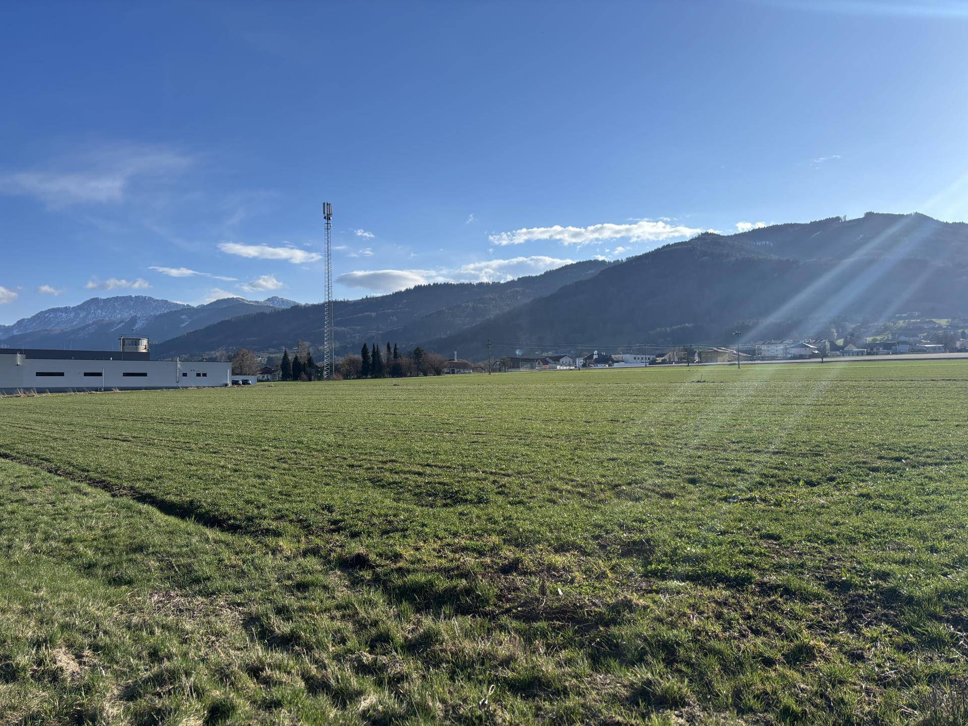 Ausgedehntes grünes Feld mit einem Sendemast, Gebäuden und majestätischen Bergen im Hintergrund bei Sonnenschein.