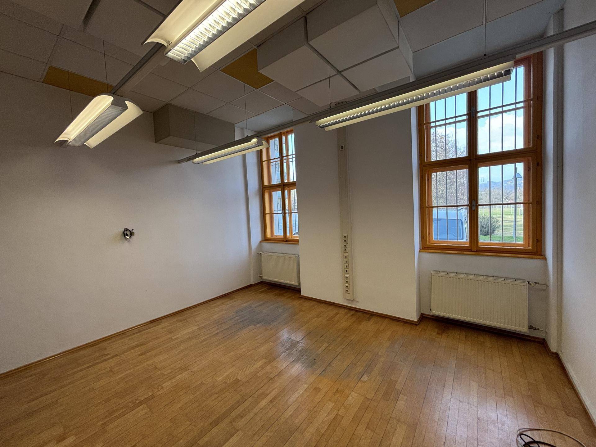 Heller Büroraum mit Holzboden, Heizkörpern unter den Fenstern und Deckenbeleuchtung, ideal für Arbeitsplätze.