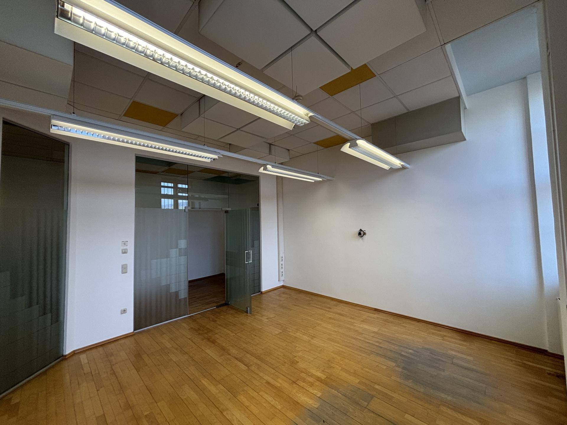 Büroraum mit Holzboden, Glaswänden und Deckenbeleuchtung, der eine moderne Arbeitsumgebung schafft.