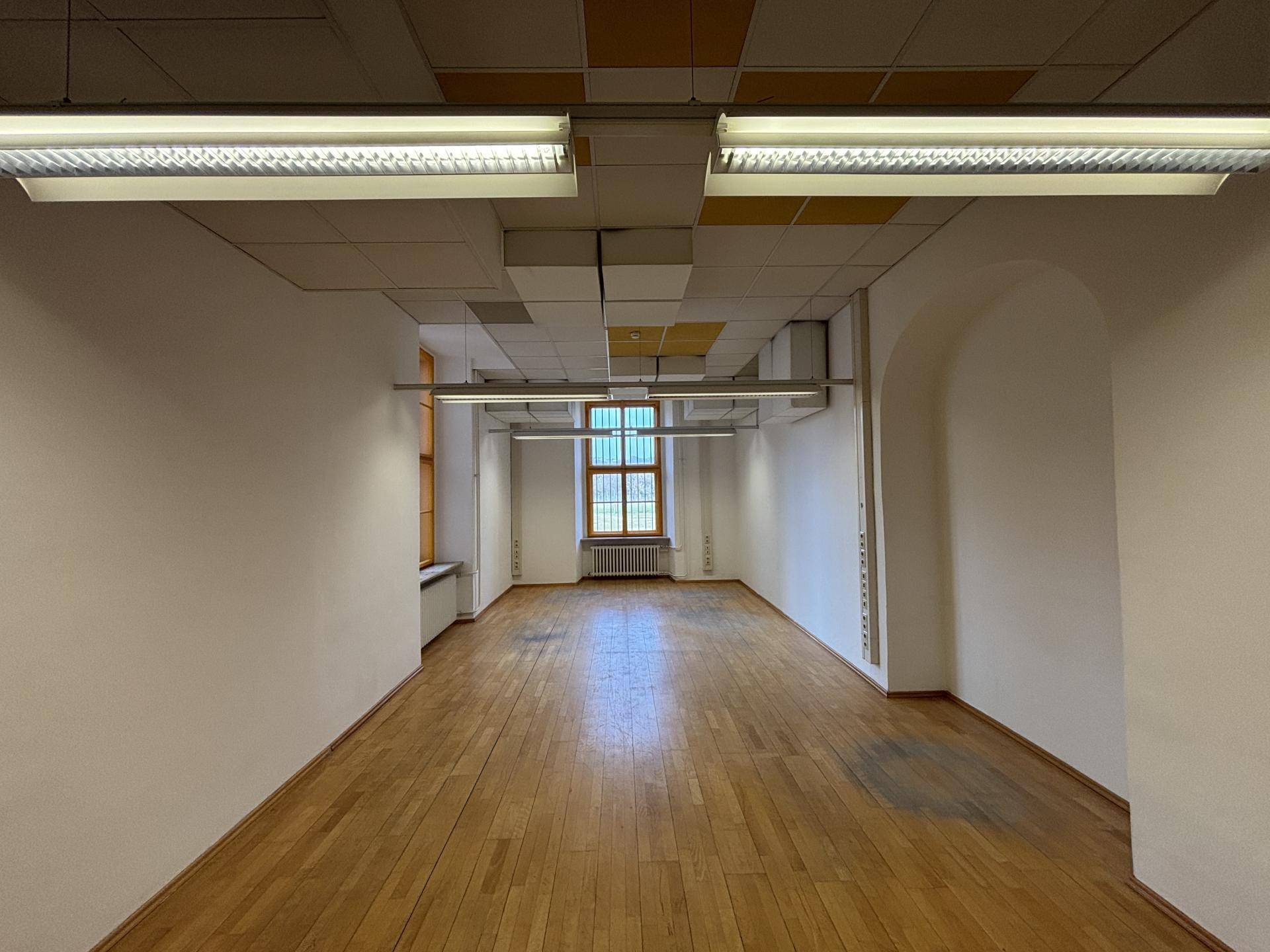 Großer, leerer Büroraum mit Holzboden, hohen Decken und Fenstern, die viel natürliches Licht hereinlassen.