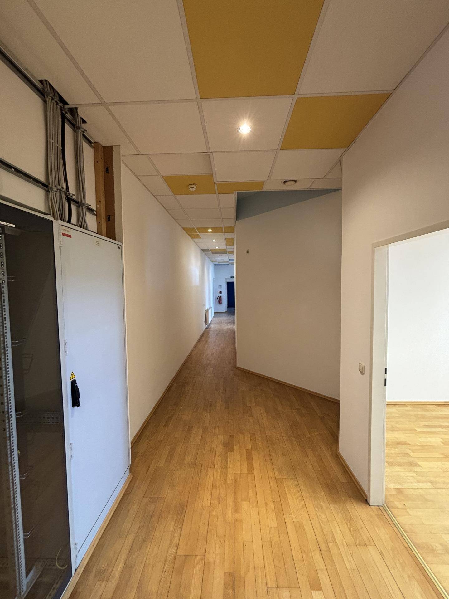 Langer Flur mit Holzboden, abgehängter Decke und sichtbaren Kabelkanälen an der Wand, führt zu weiteren Räumen.