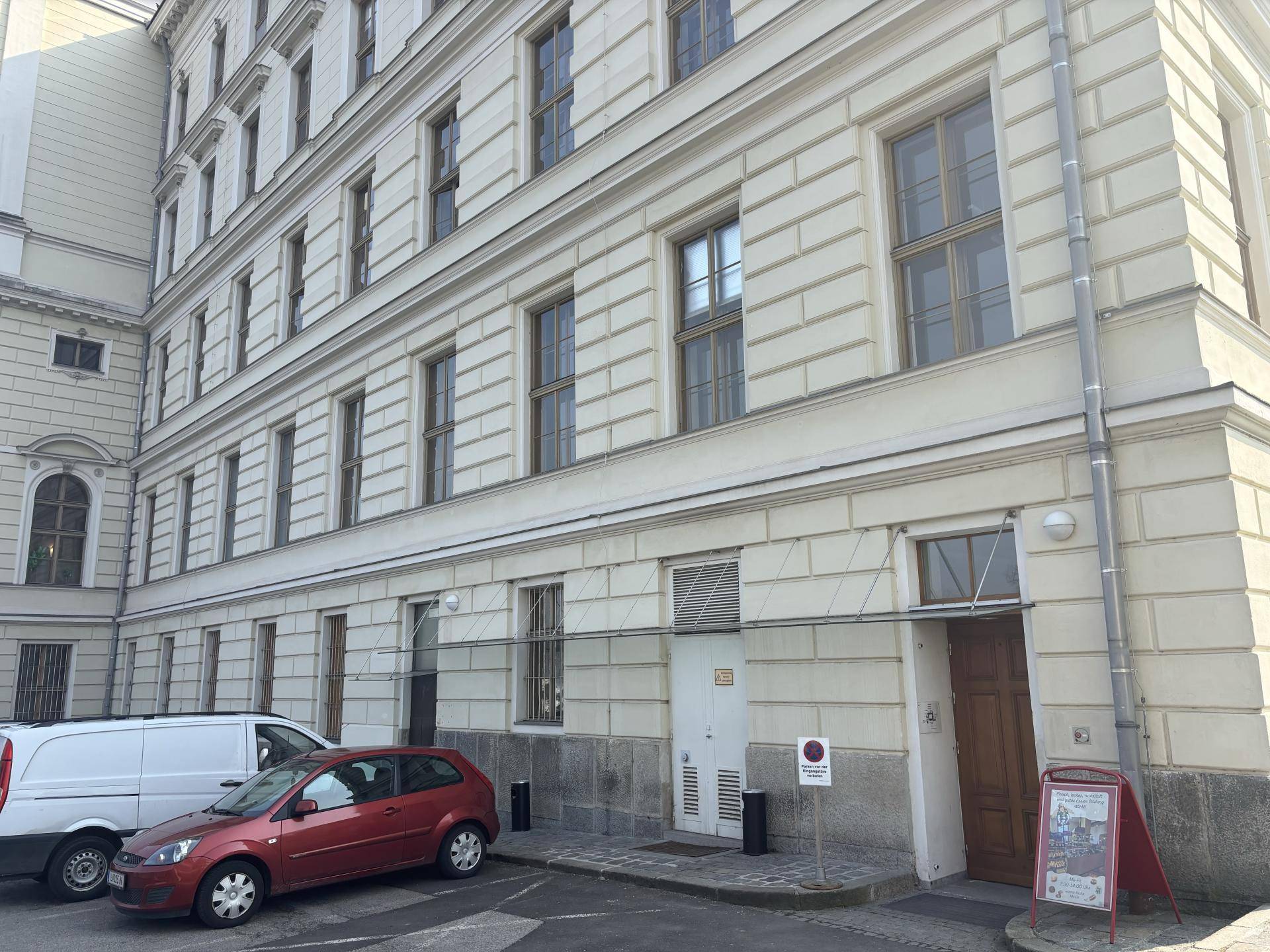 Seitenansicht eines klassischen Gebäudes mit Eingangsbereich und geparkten Autos davor, in städtischer Umgebung.