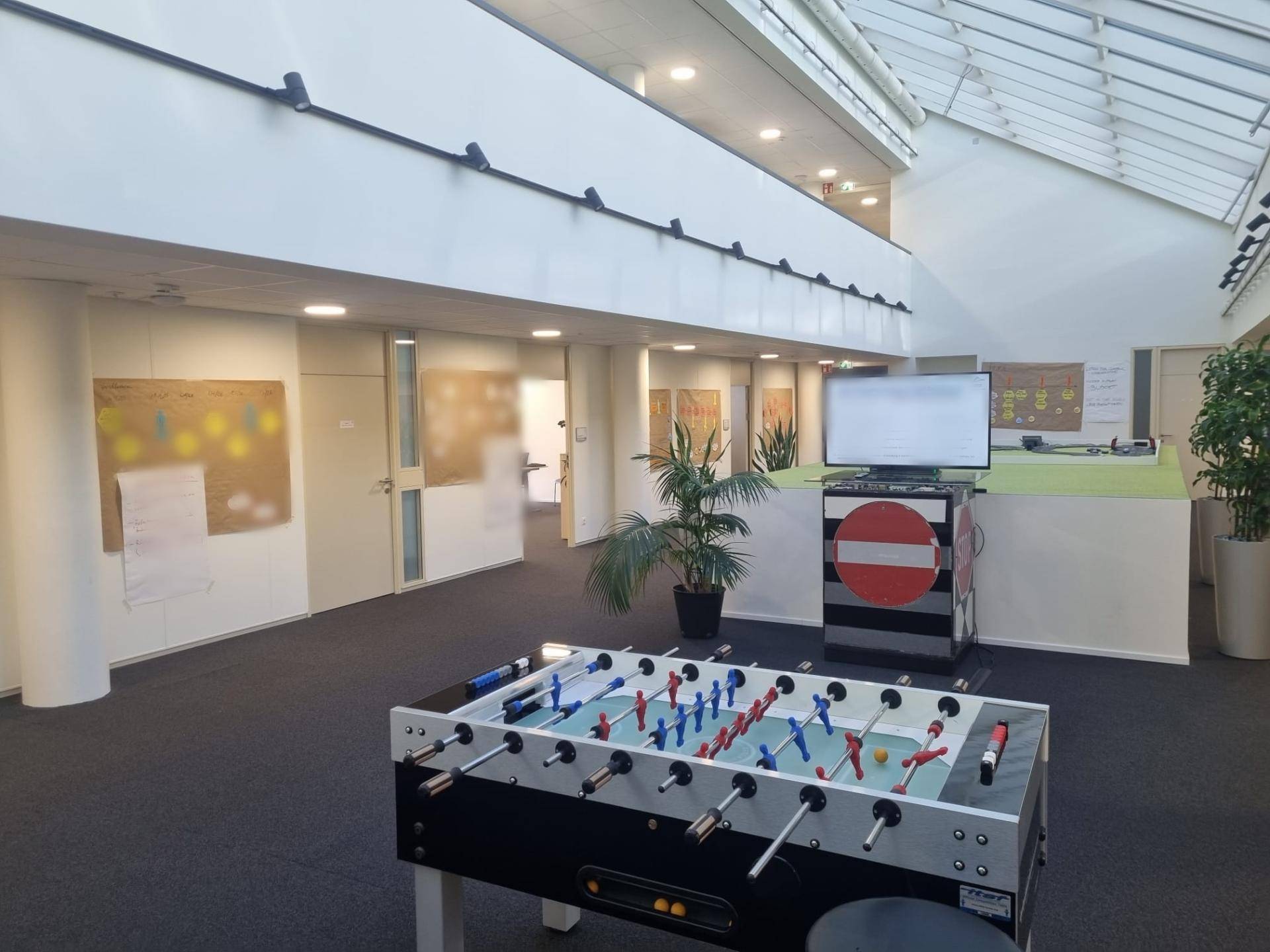 Heller Gemeinschaftsbereich in einem Bürogebäude mit Tischfußball, Oberlichtern und Pflanzen für eine entspannte Atmosphäre.