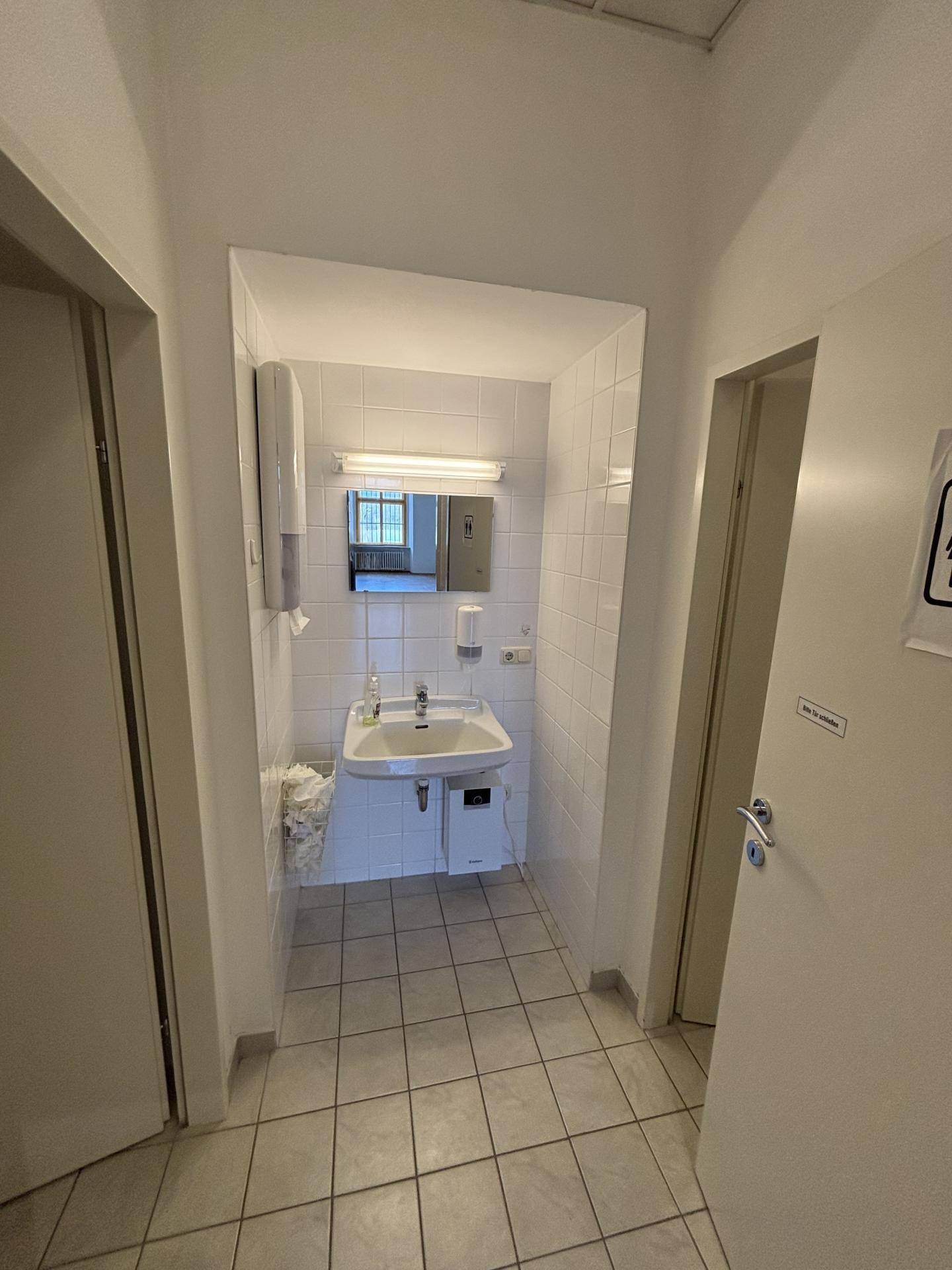 Kleiner Waschraum mit Waschbecken, Spiegel und weißen Fliesen an Wänden und Boden, ideal als Gästetoilette.
