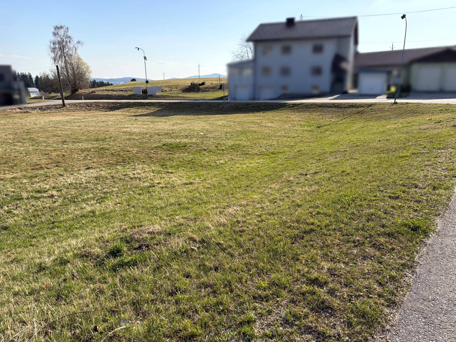 Grüne Wiesenfläche neben einer Straße mit einem Mehrfamilienhaus im Hintergrund unter blauem Himmel.