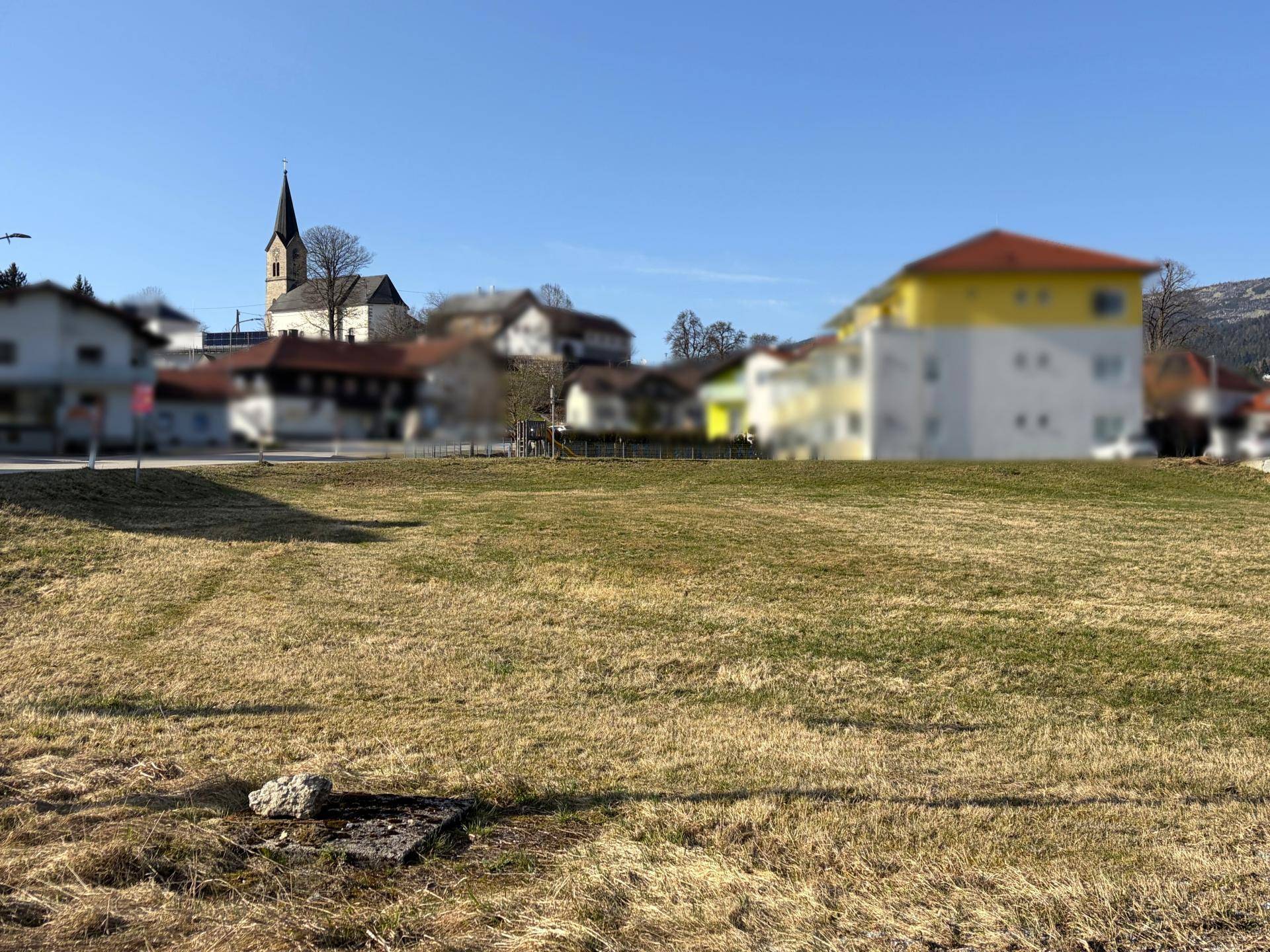 Offene Wiesenfläche mit Blick auf ein Dorf mit Kirche und Wohnhäusern im Hintergrund unter klarem Himmel.
