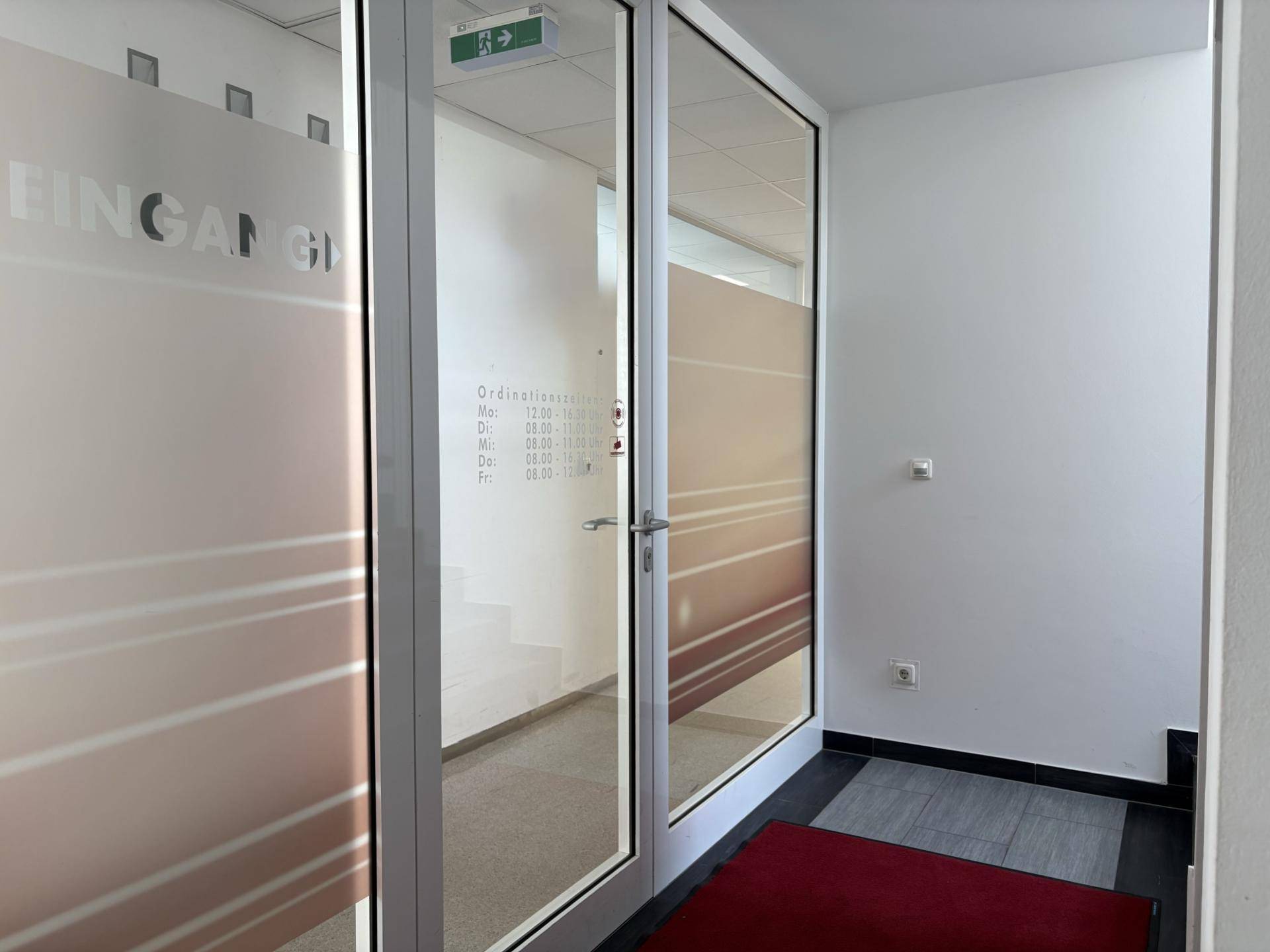 Heller Eingangsbereich mit Glastüren und Informationen zu Öffnungszeiten.