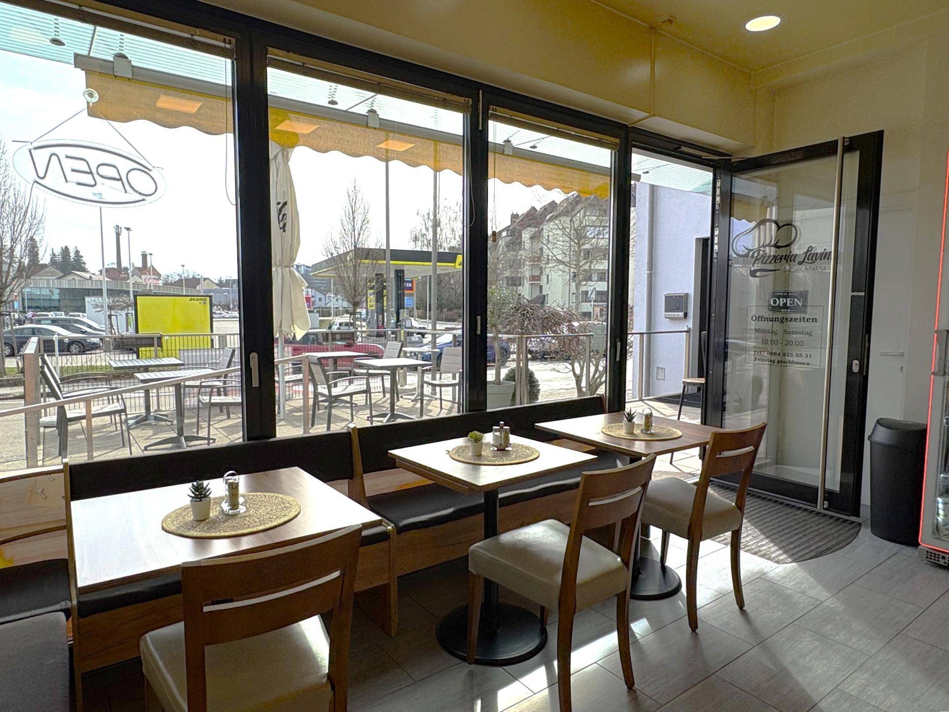 Helle Innenansicht eines Cafés mit Tischen, Stühlen und Blick auf den Außenbereich.