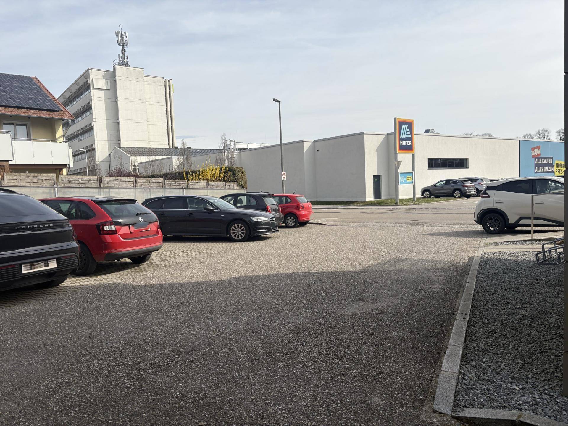 Außenparkplätze vor einem Supermarkt mit weiteren Gebäuden im Hintergrund.