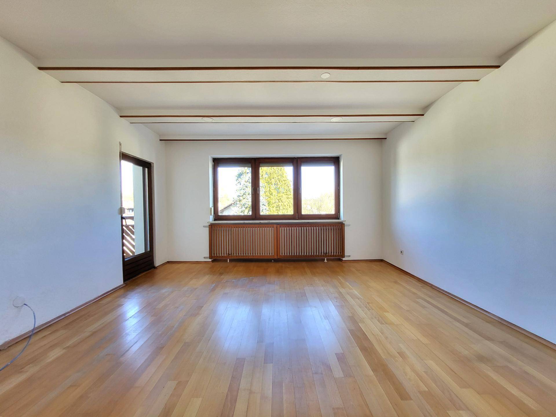 Helles, leeres Wohnzimmer mit Holzbalkendecke, Parkettboden und Zugang zum Balkon.