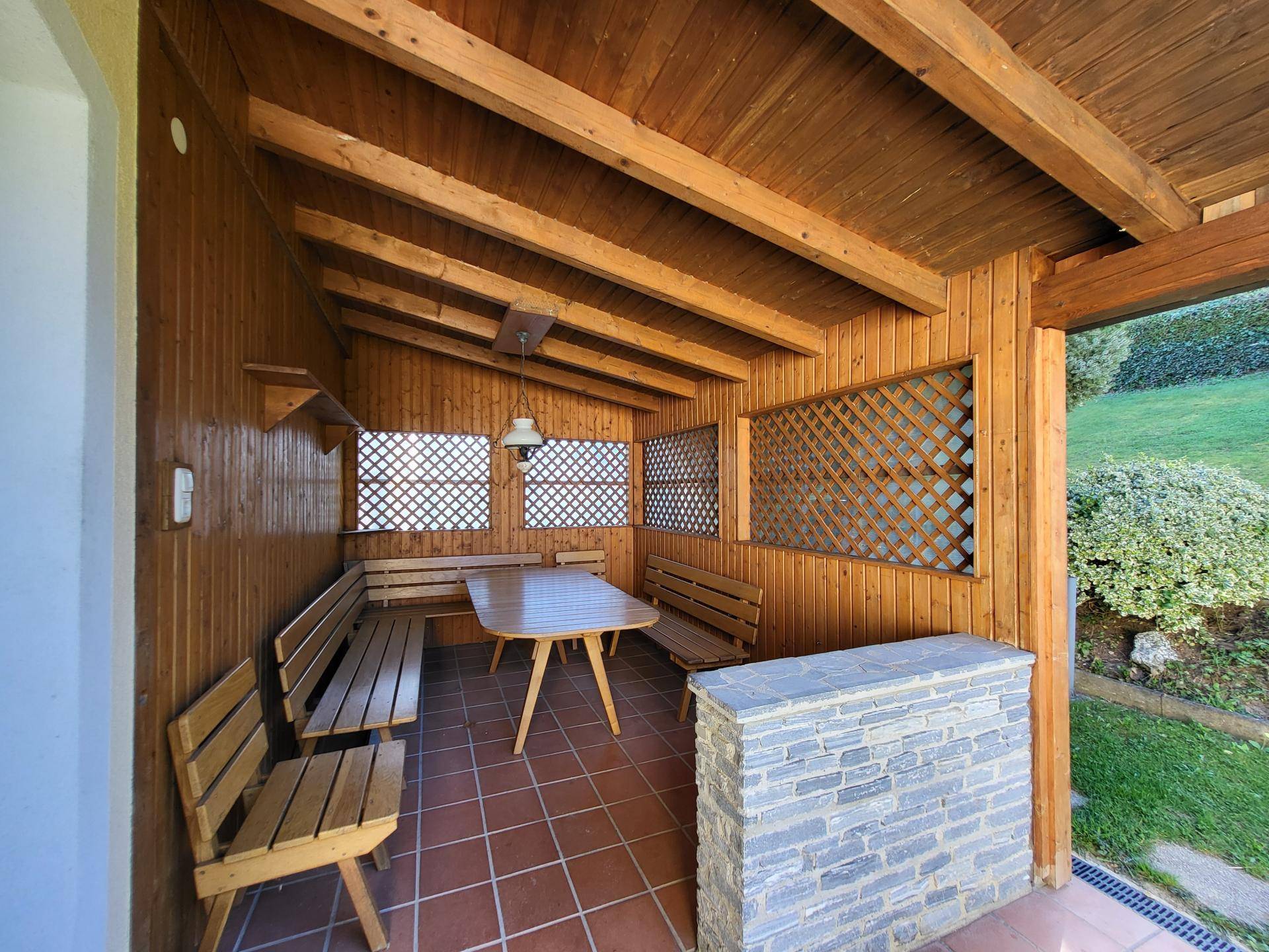Überdachte Terrasse mit Holzbänken, Tisch und Fliesenboden, ideal für gesellige Stunden.