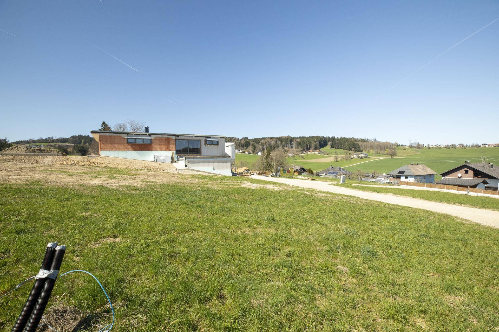 Freie Fläche eines Baugrundstücks mit einem modernen Gebäude im Hintergrund und weitem Blick ins Grüne.