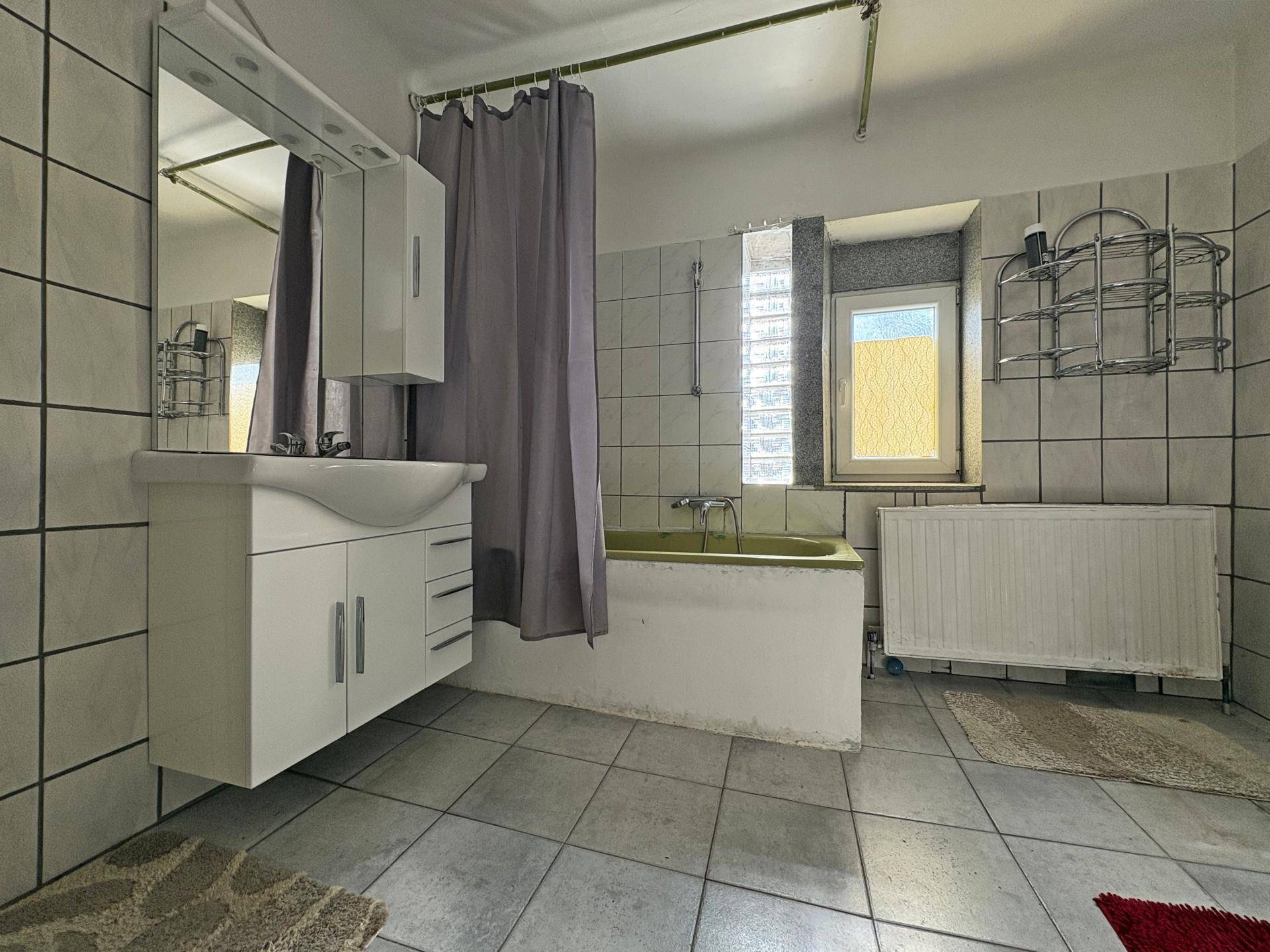 Badezimmer mit hellgrauen Fliesen, Badewanne und Waschtisch mit Spiegelschrank und Fenster.