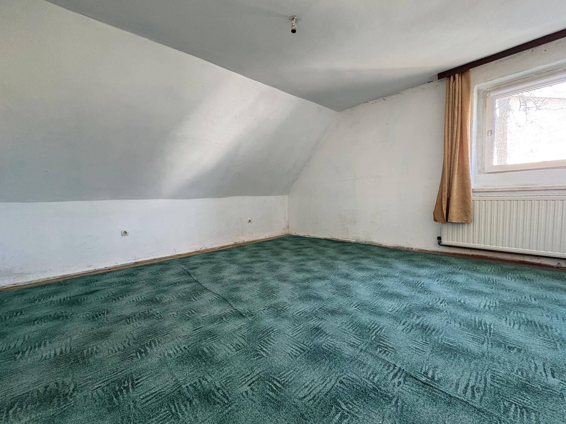 Dachzimmer mit grünem Teppichboden und schrägen Wänden, gut beleuchtet durch ein Fenster.
