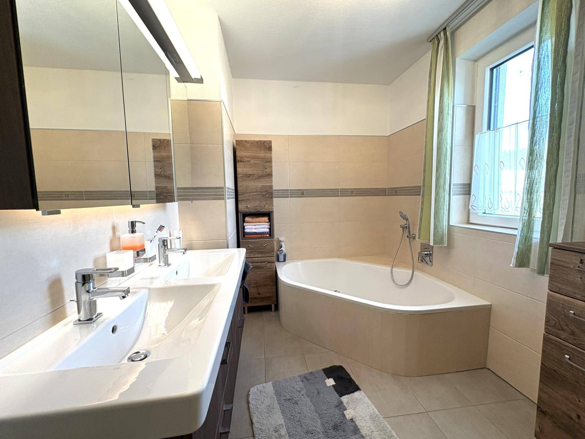 Helles Badezimmer mit Doppelwaschbecken, Eckbadewanne und Fenster für Tageslicht.