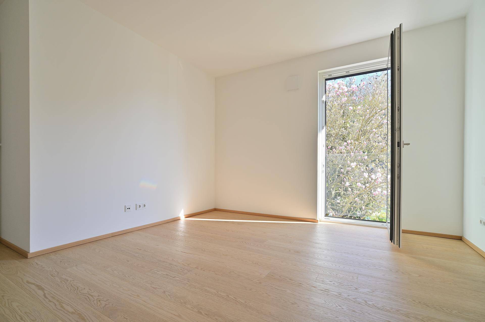 Leeres, helles Zimmer mit Holzboden und Fenster mit Blick auf blühende Bäume.