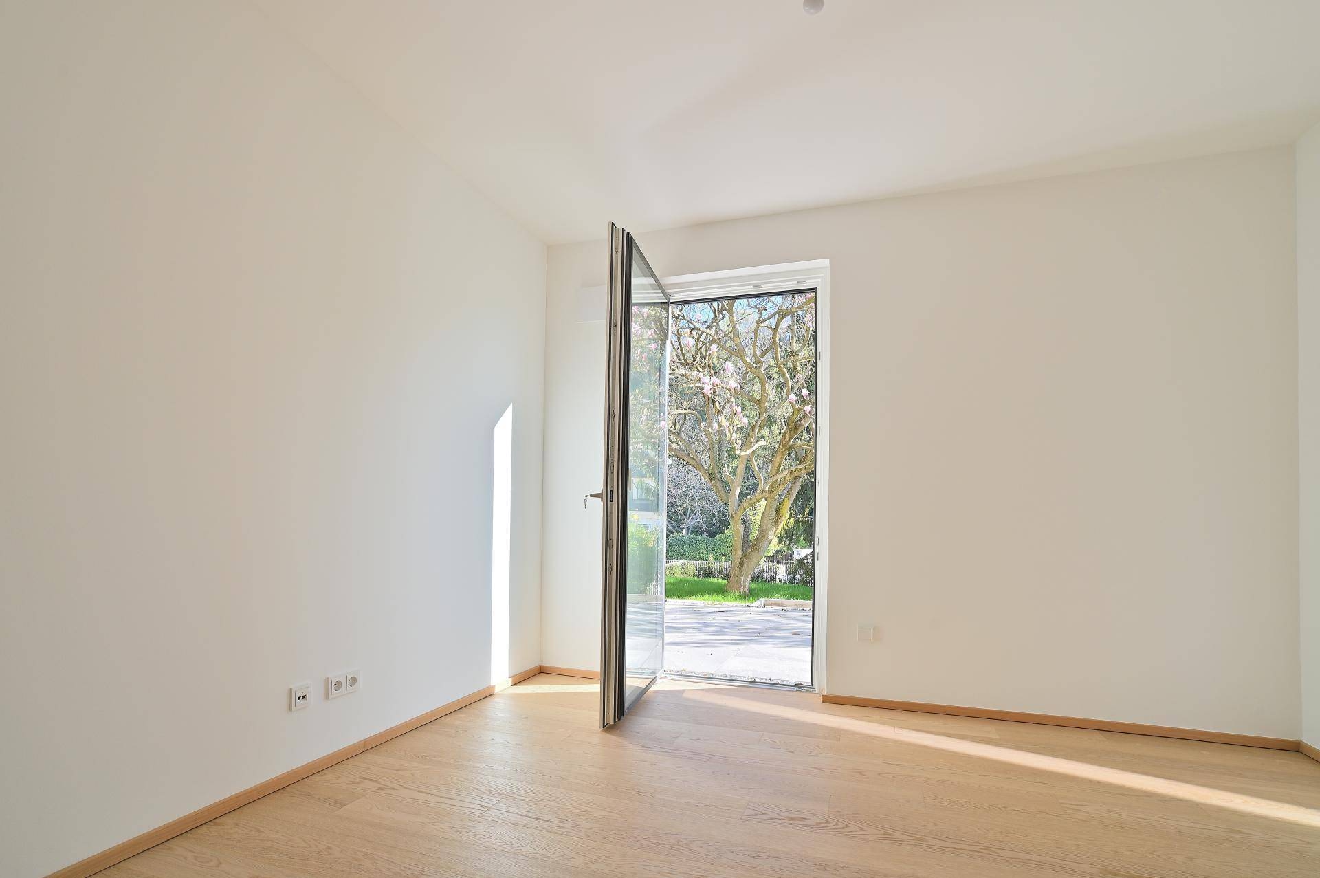 Helles, leeres Zimmer mit Holzboden und weit geöffneter Glastür zum Außenbereich.