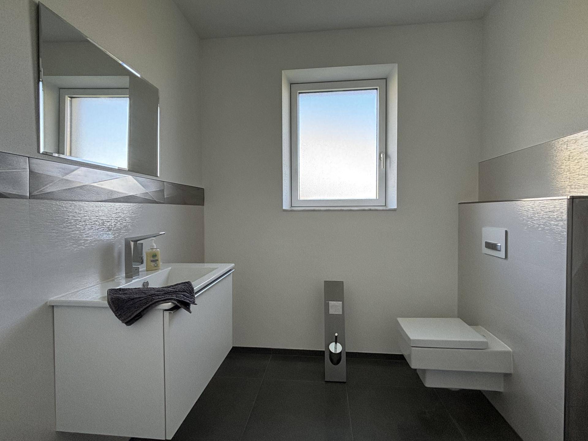 Modernes Badezimmer mit Einzelwaschbecken, wandhängendem WC und Fenster, in hellen Grautönen gehalten.