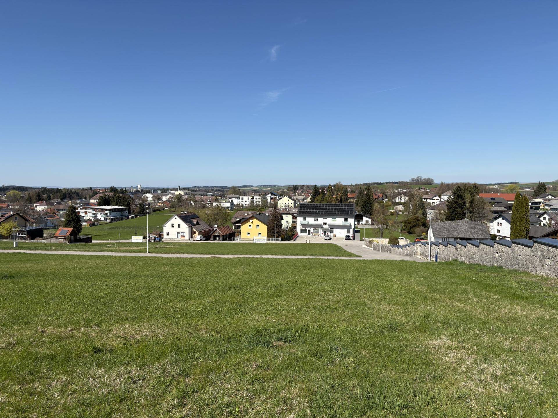 Panoramablick über eine weite Wiese auf eine Siedlung mit verschiedenen Häusern und klarem Himmel.