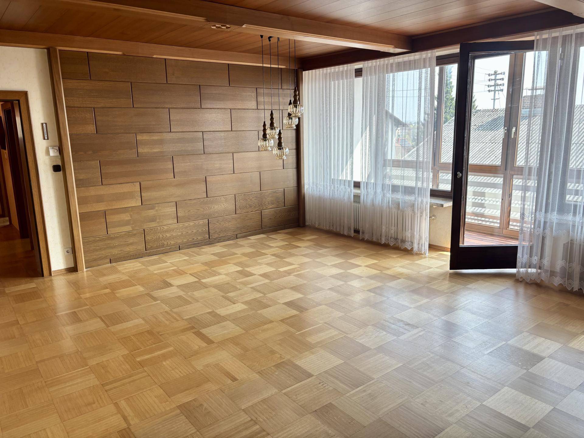 Geräumiges Wohnzimmer mit Holzwandverkleidung, Parkettboden und Zugang zum Balkon.