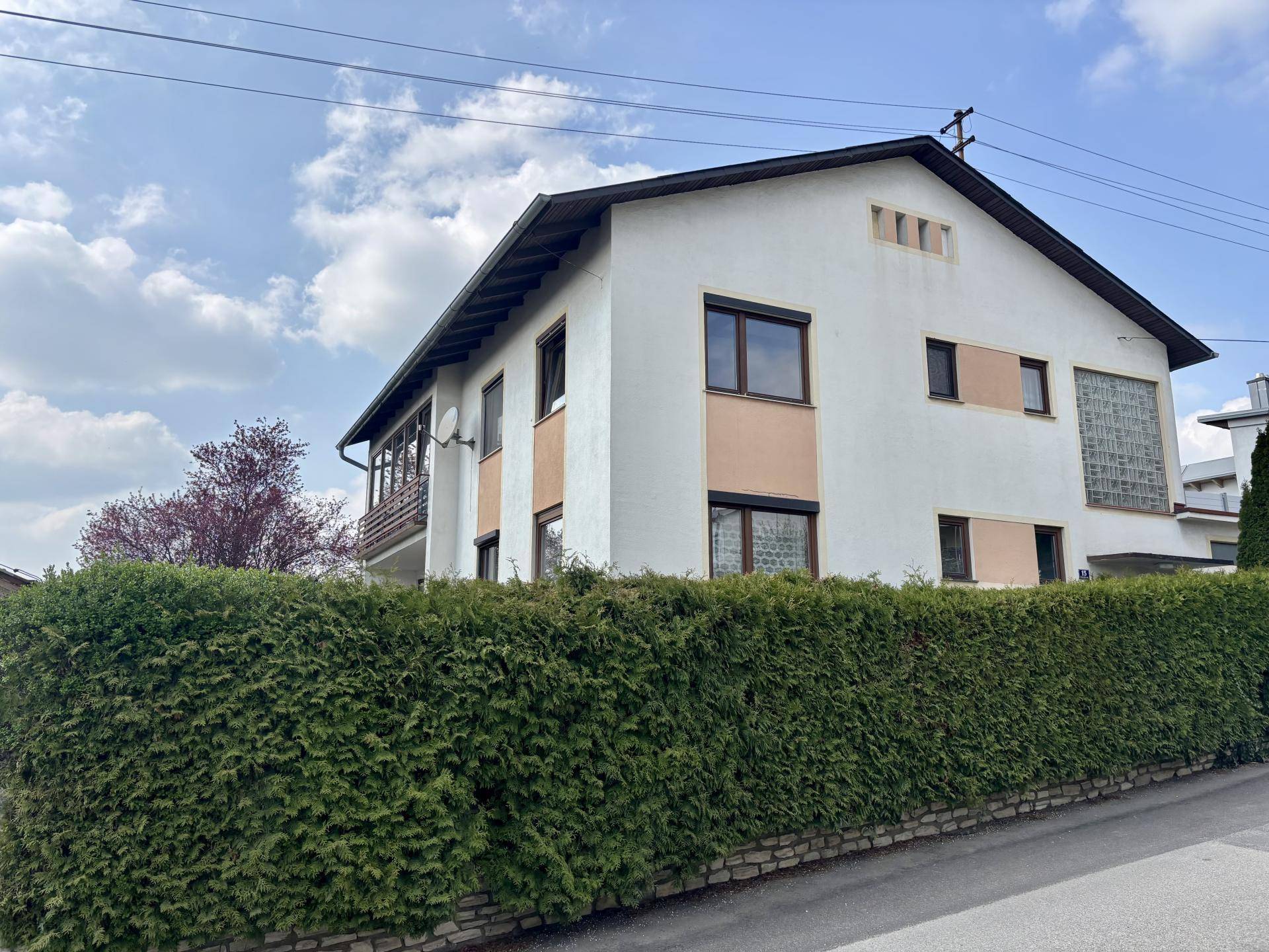 Seitenansicht des Hauses mit gepflegter Hecke, Balkon und Satteldach unter blauem Himmel.