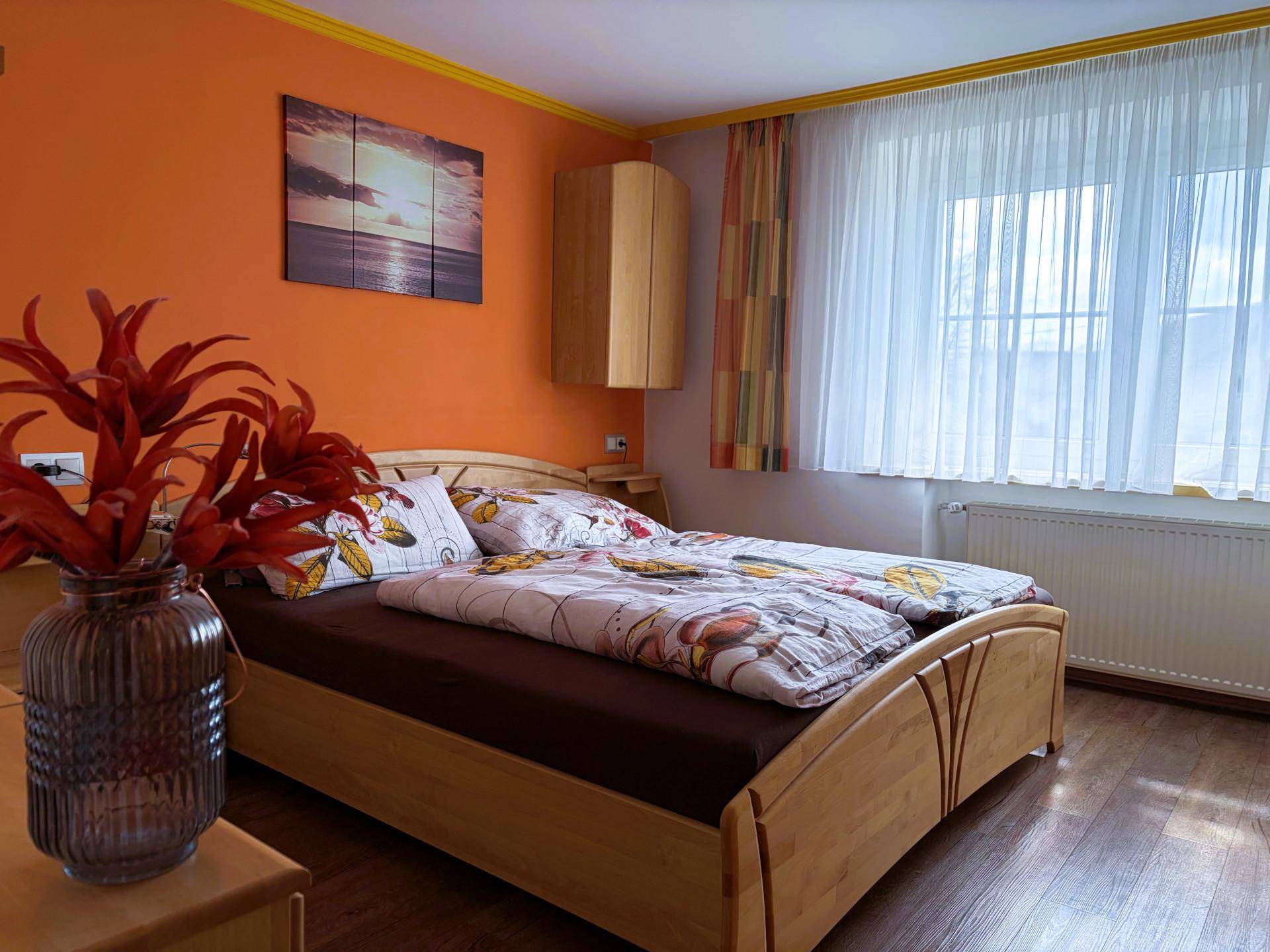 Schlafzimmer mit orangefarbenen Wänden, hellem Holzdoppelbett und großem Fenster.