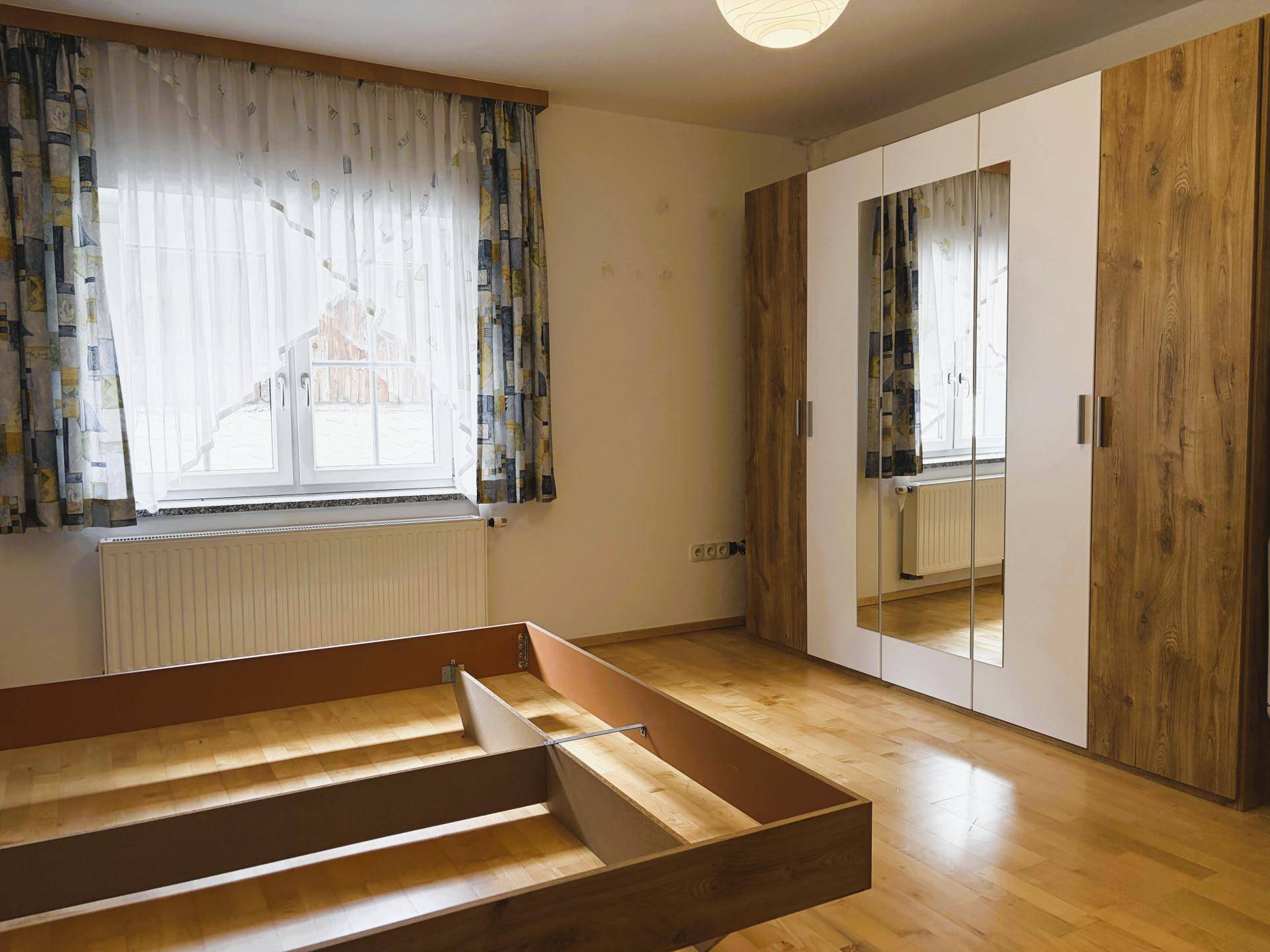 Helles Schlafzimmer mit Holzboden, Spiegelschrank und Fenster mit Blick nach draußen.