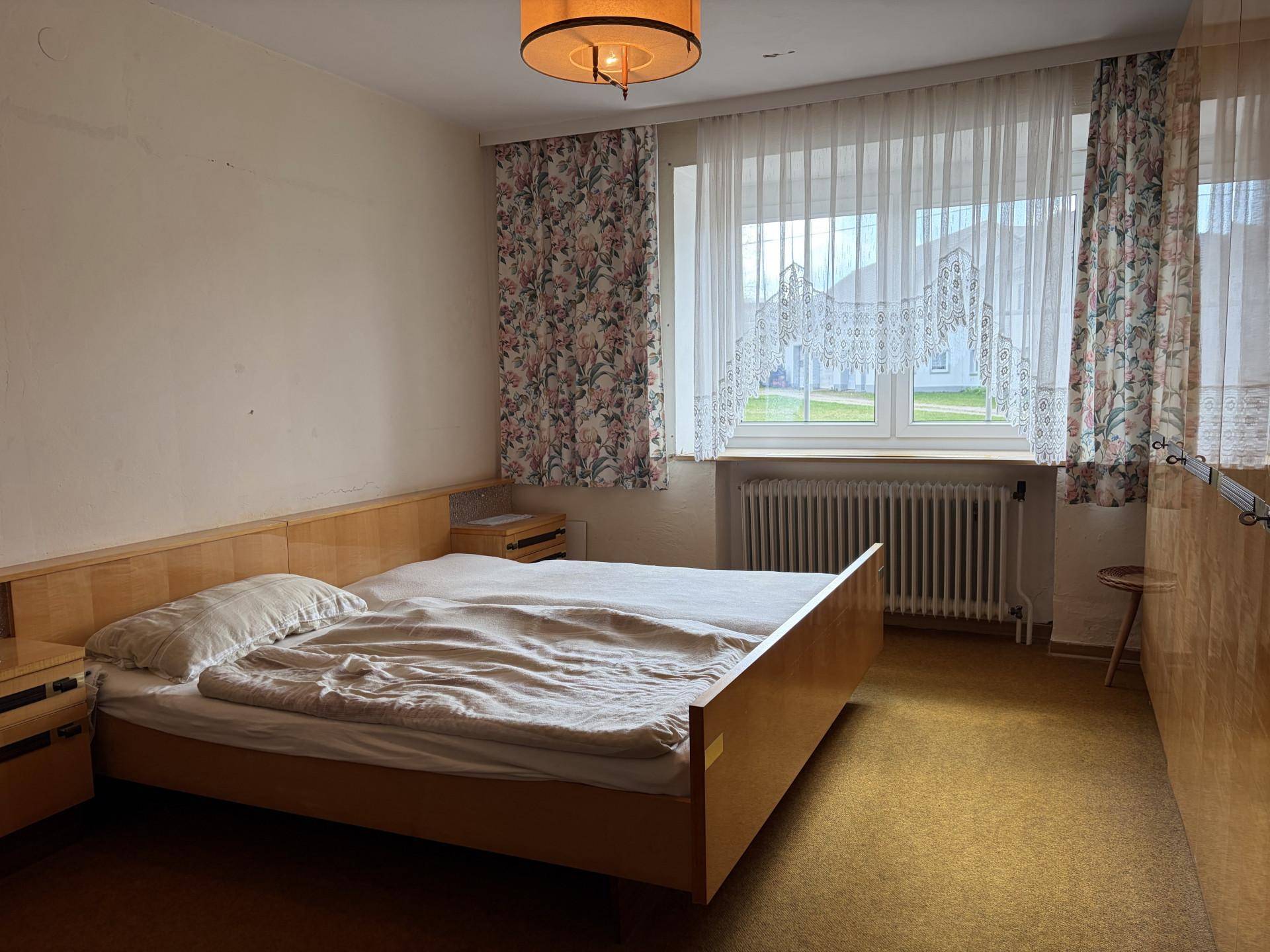 Schlafzimmer mit Doppelbett, Teppichboden und Fenster mit floralen Vorhängen.