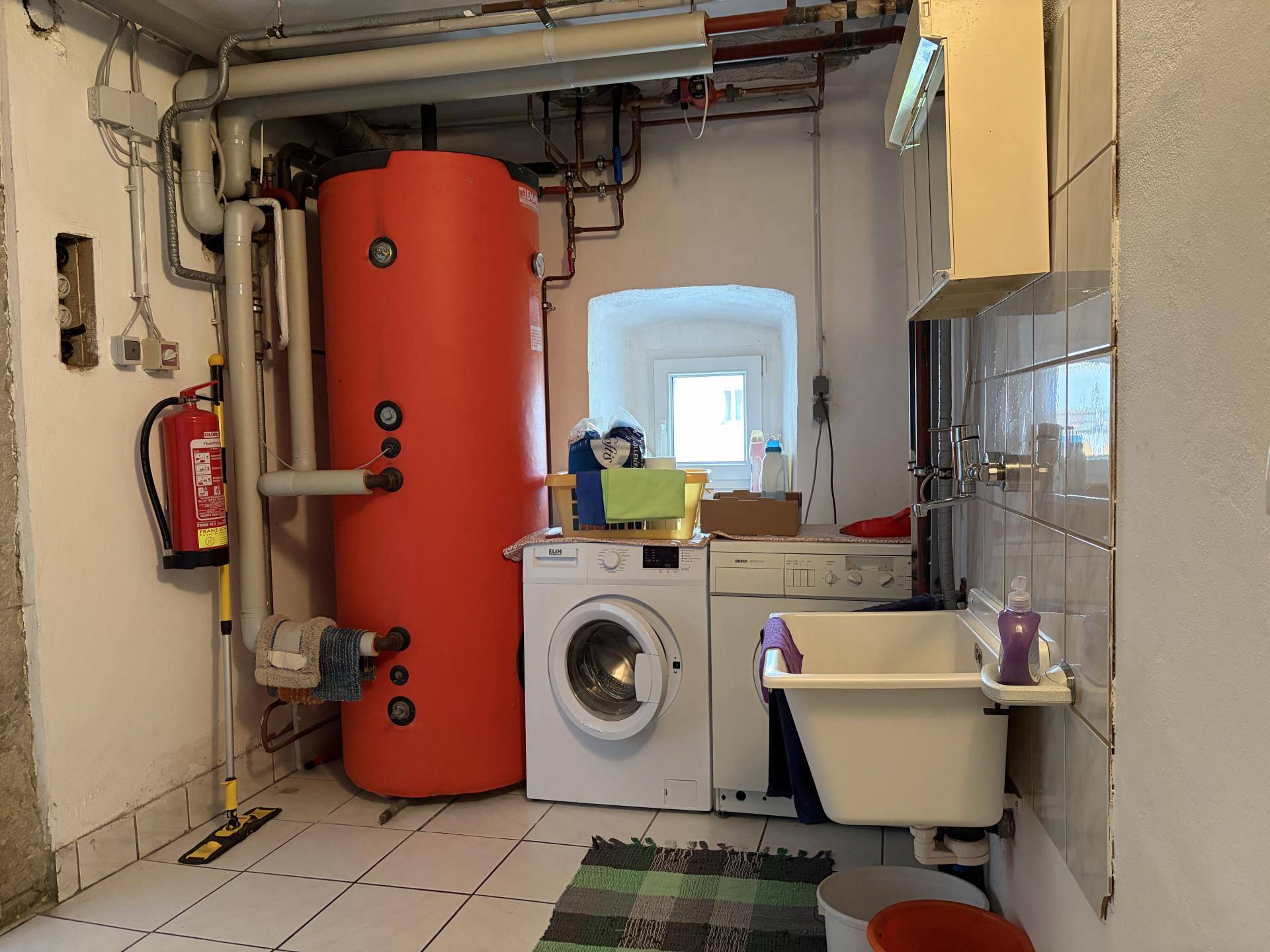 Hauswirtschaftsraum im Keller mit Waschmaschine, Warmwasserspeicher und kleinem Fenster.
