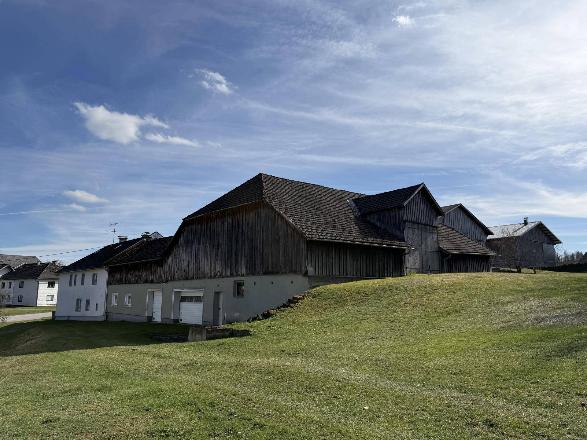 Langes Scheunengebäude mit Holzwänden und Satteldach, eingebettet in eine grüne Hügellandschaft.