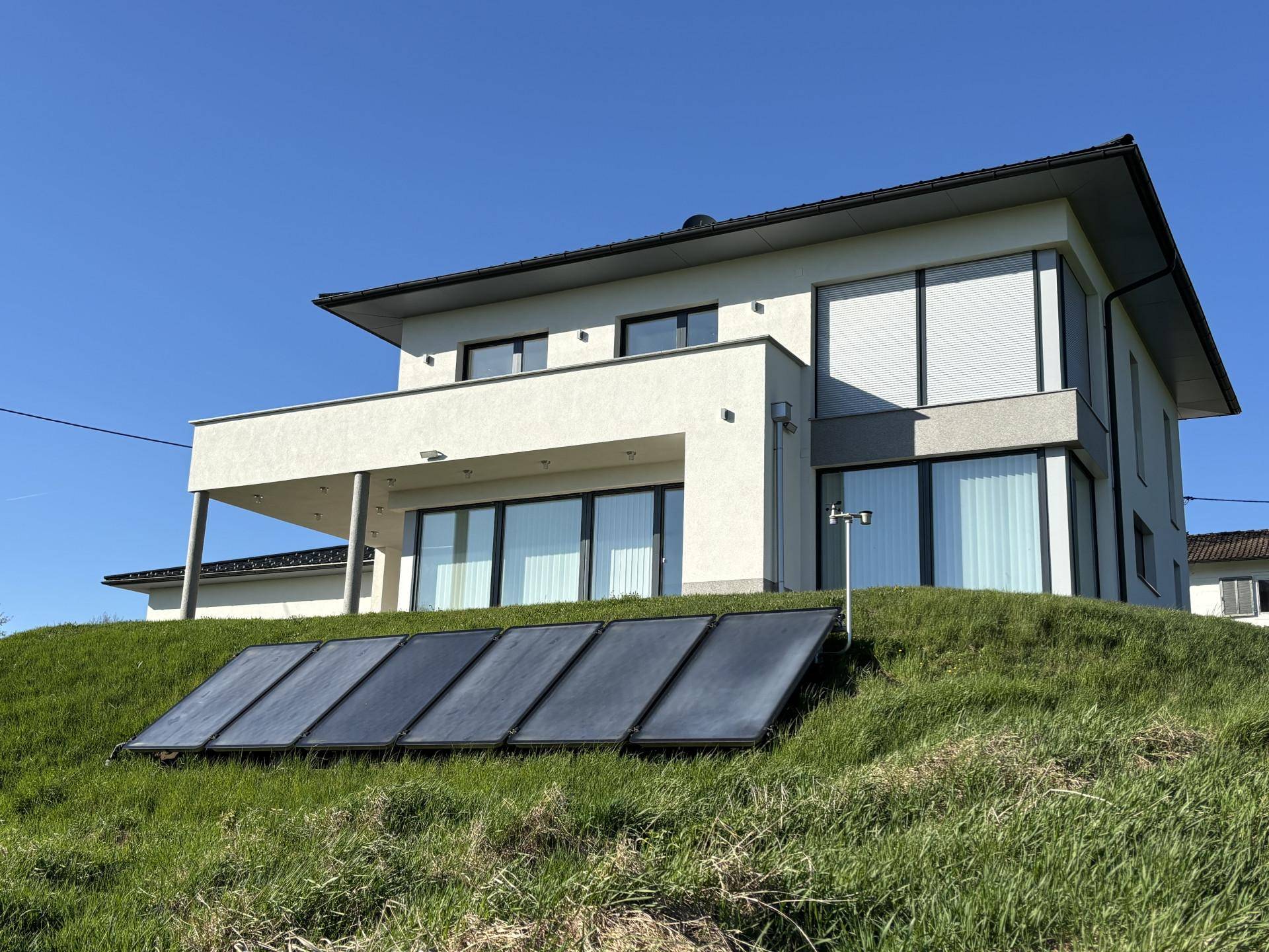 Modernes Haus mit Photovoltaikanlage auf dem grünen Hang im Vordergrund, unter blauem Himmel.
