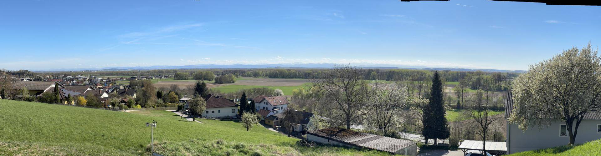 Weitläufiges Panorama der ländlichen Umgebung mit Dorf, Feldern und einer Bergkette am Horizont.