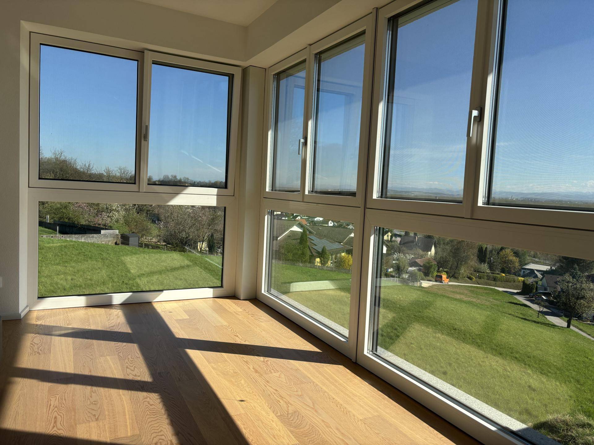 Helles Zimmer mit großen Eckfenstern und Holzboden, bietet einen weiten Panoramablick ins Grüne.