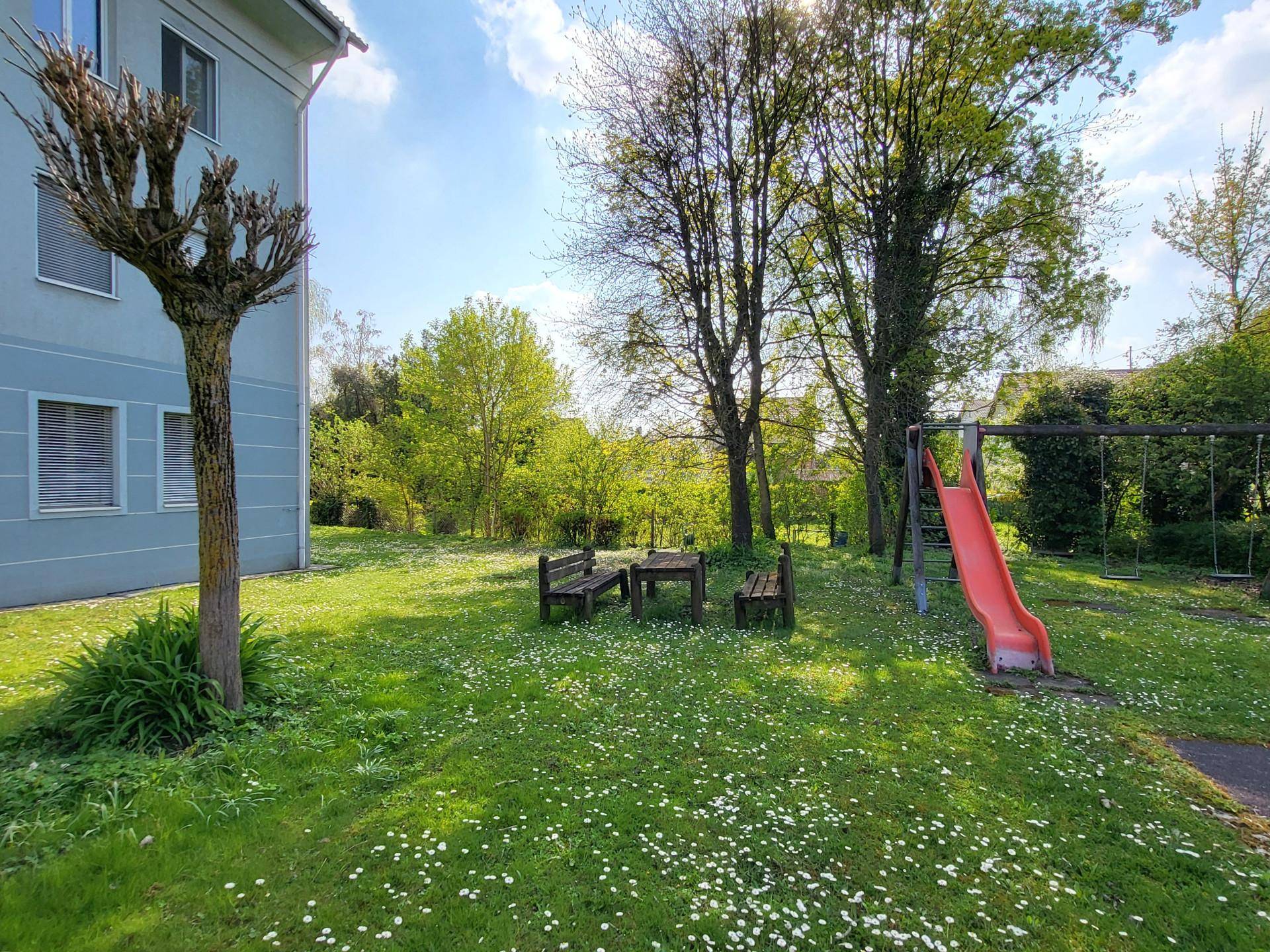 Grüner Gemeinschaftsgarten mit Spielplatz, Rutsche und Sitzgelegenheiten im Freien.