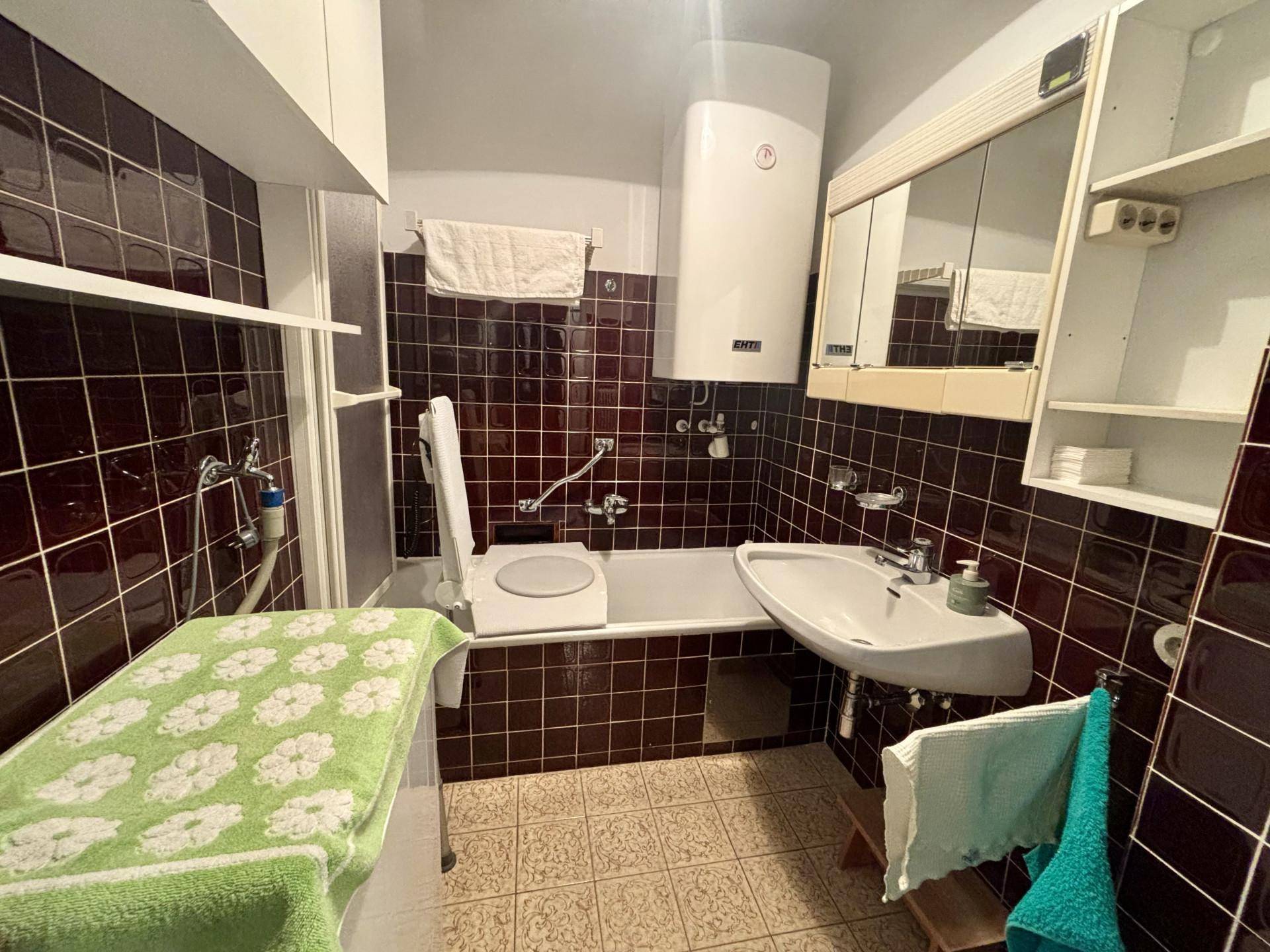 Badezimmer im 80er-Jahre-Stil mit dunklen Fliesen, Badewanne und Einzelwaschbecken.