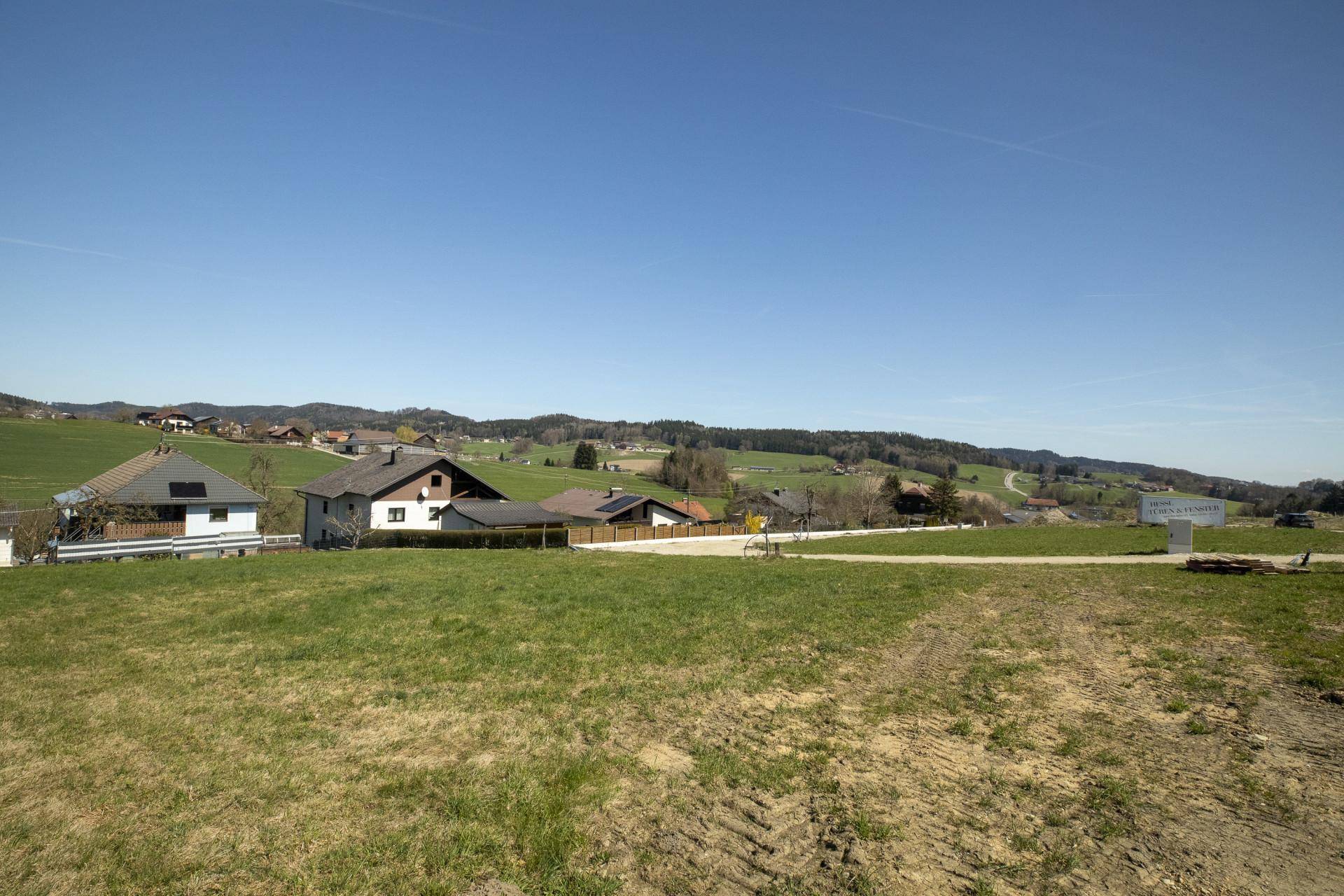 Offenes Baugrundstück mit weitem Blick über die grüne Landschaft und einige Wohnhäuser am Horizont.