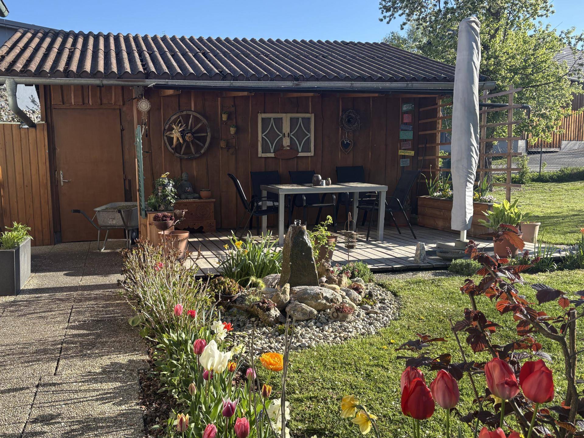 Sonniger Gartenbereich mit Steingarten, Blumen und überdachtem Essplatz.